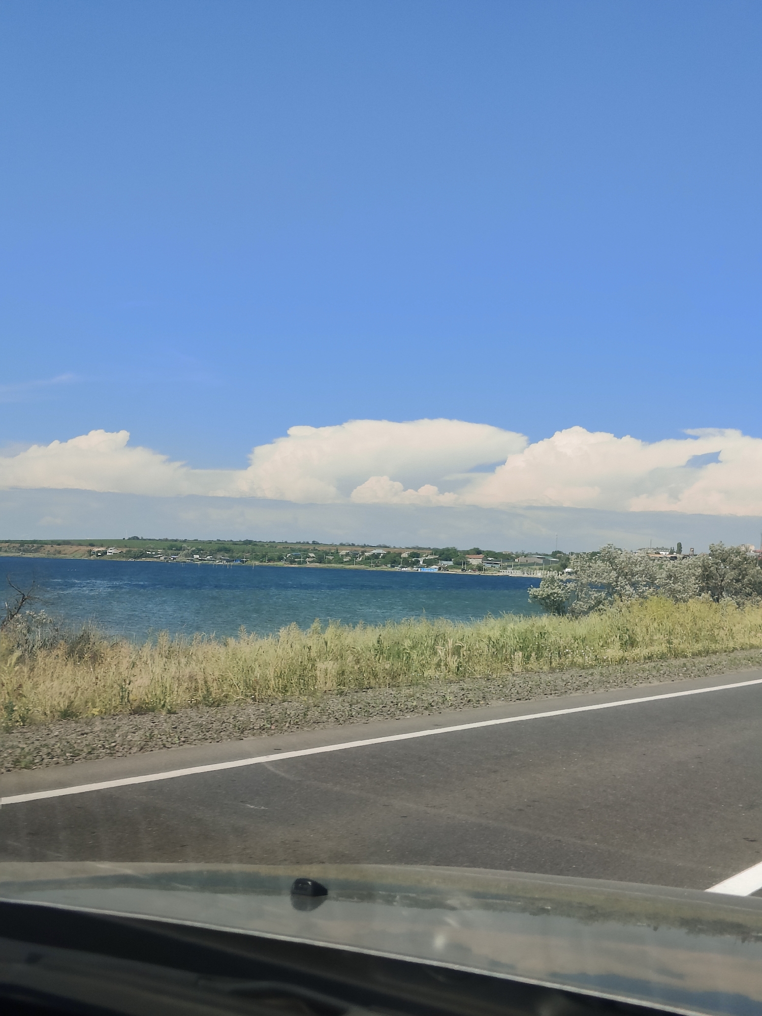 The image shows a scenic view of a body of water with a town in the distance, seen from a road. The sky is blue with fluffy white clouds. The foreground features a paved road with a white line marking the edge. To the left of the road, there is a strip of gravel and then tall, dry grass. Beyond the grass is the blue water, which appears to be a bay or lake. In the background, across the water, is a line of buildings and trees, indicating a town or village. The overall environment suggests a warm, clear day in a coastal or lakeside area. It is a still scene, with no people or animals visible, and no apparent action taking place. The perspective suggests the photo was taken from a moving vehicle.