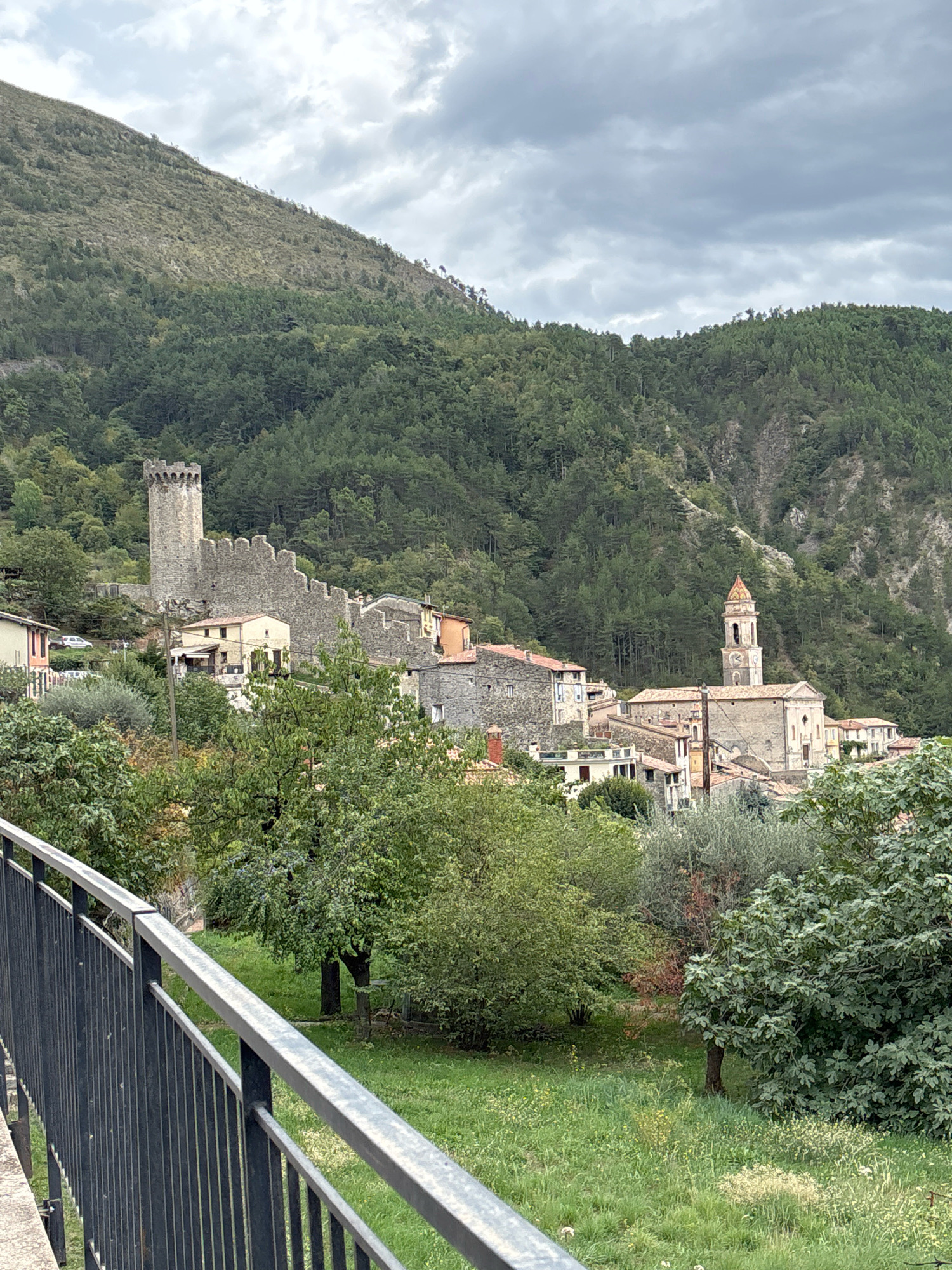 The image depicts a quaint village nestled in a valley, with a stone castle ruin on a hill overlooking the settlement. The castle features a prominent square tower and crenellated walls. Below the castle, a cluster of stone buildings with red-tiled roofs forms the village. A church with a bell tower topped with a colorful, patterned roof stands in the center of the village. The surrounding landscape is characterized by steep, tree-covered mountains and a cloudy sky. In the foreground, a dark metal railing of a bridge or walkway is visible, separating the viewer from a grassy area with scattered trees. The scene is peaceful and evokes a sense of historical charm and rural tranquility.