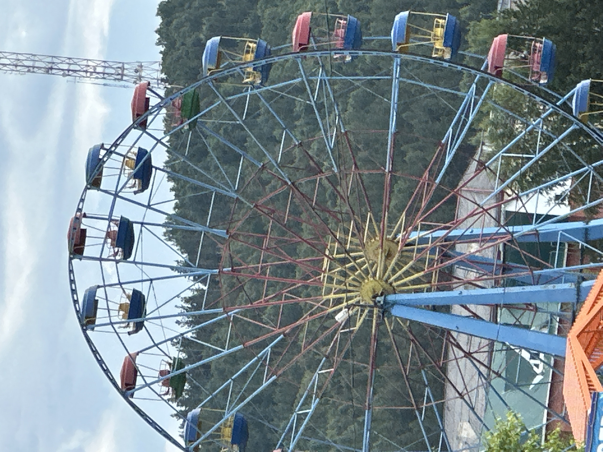A Ferris wheel with blue and red cabins is seen against a backdrop of a lush green hillside and a cloudy sky. A tall metal tower, possibly for communication, is visible in the upper right quadrant of the image. The Ferris wheel is colorful, with blue, red, and yellow accents on its cabins. The structure appears to be made of metal, with a central yellow hub and numerous spokes radiating outwards. Some of the cabins are occupied by people, though their faces are not clearly discernible. The overall scene suggests an amusement park or recreational area situated in a natural setting.