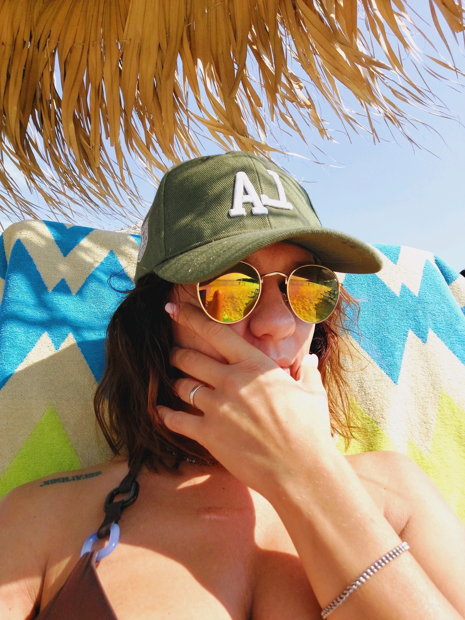 A person is lying down under a straw umbrella on a beach in Sozopol, Bulgaria, holding their hand to their face, partially obscuring their sunglasses and nose. They are wearing a green baseball cap with "AJ" embroidered on it in white. The person is also wearing a bikini and a bracelet on their arm. A towel with a blue and yellow zig-zag pattern is behind them. The sky is visible above the umbrella, and the sunlight is bright, creating shadows on the person's body. The reflection in the sunglasses shows a beach scene with lounge chairs and umbrellas.