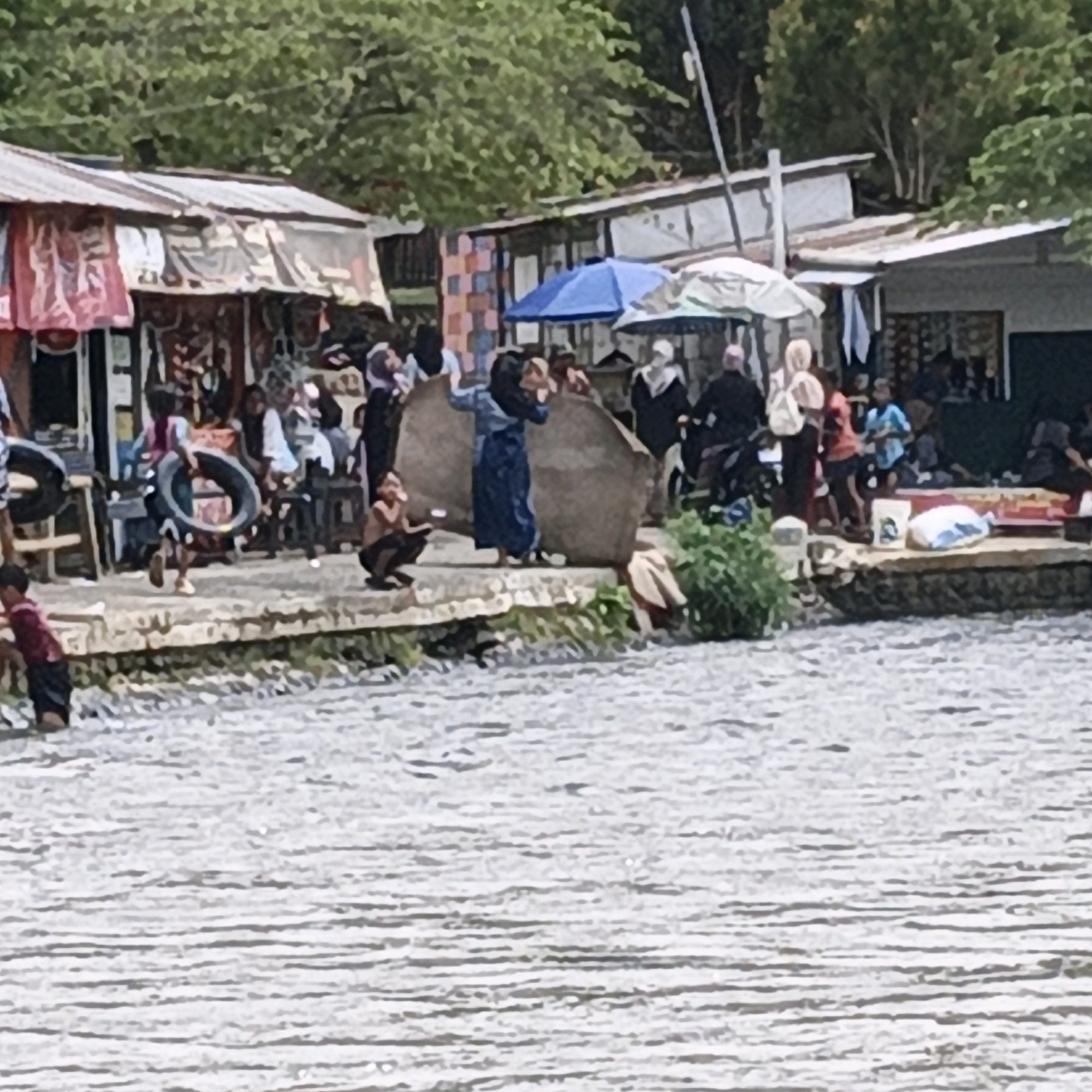 In Penusupan, Indonesia, a busy waterfront scene unfolds with various individuals and activities. Along the edge of a body of water, a row of stalls with corrugated metal roofs are set up. These stalls display various items, some with red and white fabric banners. A blue and white umbrella is visible between two stalls, suggesting a marketplace or gathering spot.
Several people are present, some are standing near the stalls, while others are closer to the water. One child in a red shirt is standing by the water's edge. Another child is running with a tire on the stone embankment separating the stalls from the water. Further down, a person in a blue dress stands near a large, round, tan object. A person wearing a black headscarf is seated near a pile of what appear to be fishing nets. Other individuals in the background are dressed in traditional clothing, including headscarves and long garments. A few people are visible in the water, appearing to be children. Green foliage can be seen behind the stalls, and a utility pole is present on the right side of the image. The water itself is choppy with visible ripples.
