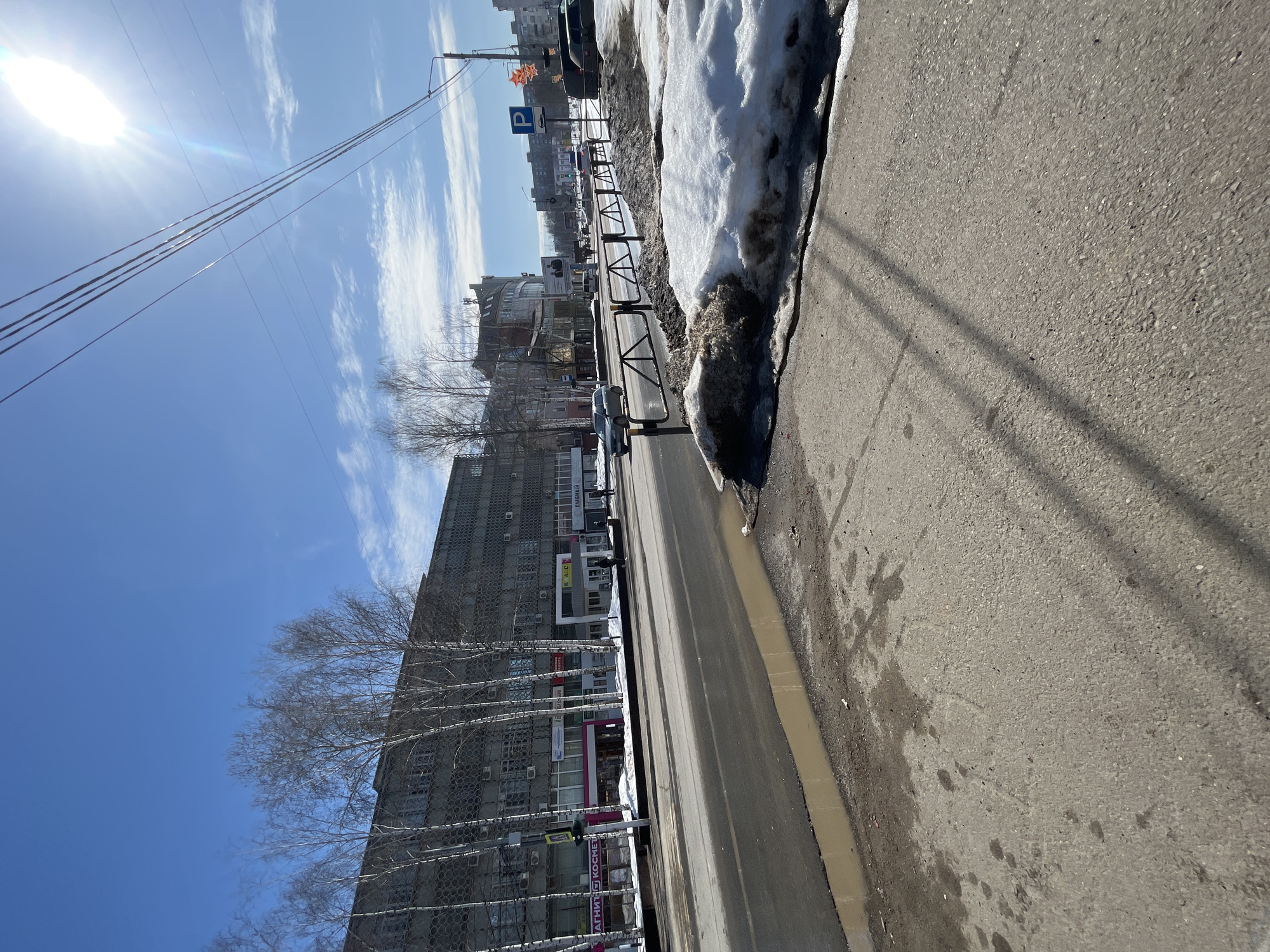 The image presents an urban street scene on Sovetskaya Ulitsa, 40, in Chaikovsky, Russia, observed under clear daylight with a bright blue sky and scattered wispy clouds. A pronounced sun flare is visible in the upper right quadrant.
The foreground consists of a grey concrete sidewalk, showing dark stains and wet patches. Shadows from overhead utility lines extend across this surface from the lower left towards the mid-right. Along the right edge of the sidewalk, accumulated piles of dirty, melting snow are present, separated from the street by a black metal pedestrian barrier.
The midground features a two-lane asphalt road, partially wet and exhibiting a large, muddy puddle at the curb in the lower left. A silver-grey sedan is seen traveling away from the camera in the left lane. Further down the road, other vehicles are visible, and a blue rectangular sign with a white 'P' (indicating parking) stands on the right side of the street, behind the snow piles.
On the left side of the street, a multi-story grey building with a patterned facade, possibly composed of pre-fabricated panels with a repeating circular or hexagonal motif, dominates the background. Several bare birch trees with white bark are positioned along the sidewalk in front of this building. The ground floor contains various retail establishments, identified by signs featuring Russian text, including "МАГНИТ КОСМЕТИК" (Magnit Kosmetik), "К.А.Л.С." (K.A.L.S.), and "РЫБНЫЙ" (Rybnyy). At least three individuals are visible on the sidewalk in front of these shops, one positioned closer to the camera and two further down. Utility poles and multiple overhead power lines are visible against the sky, particularly in the upper right. The overall environment, characterized by dirty, melting snow, suggests a late winter or early spring season.