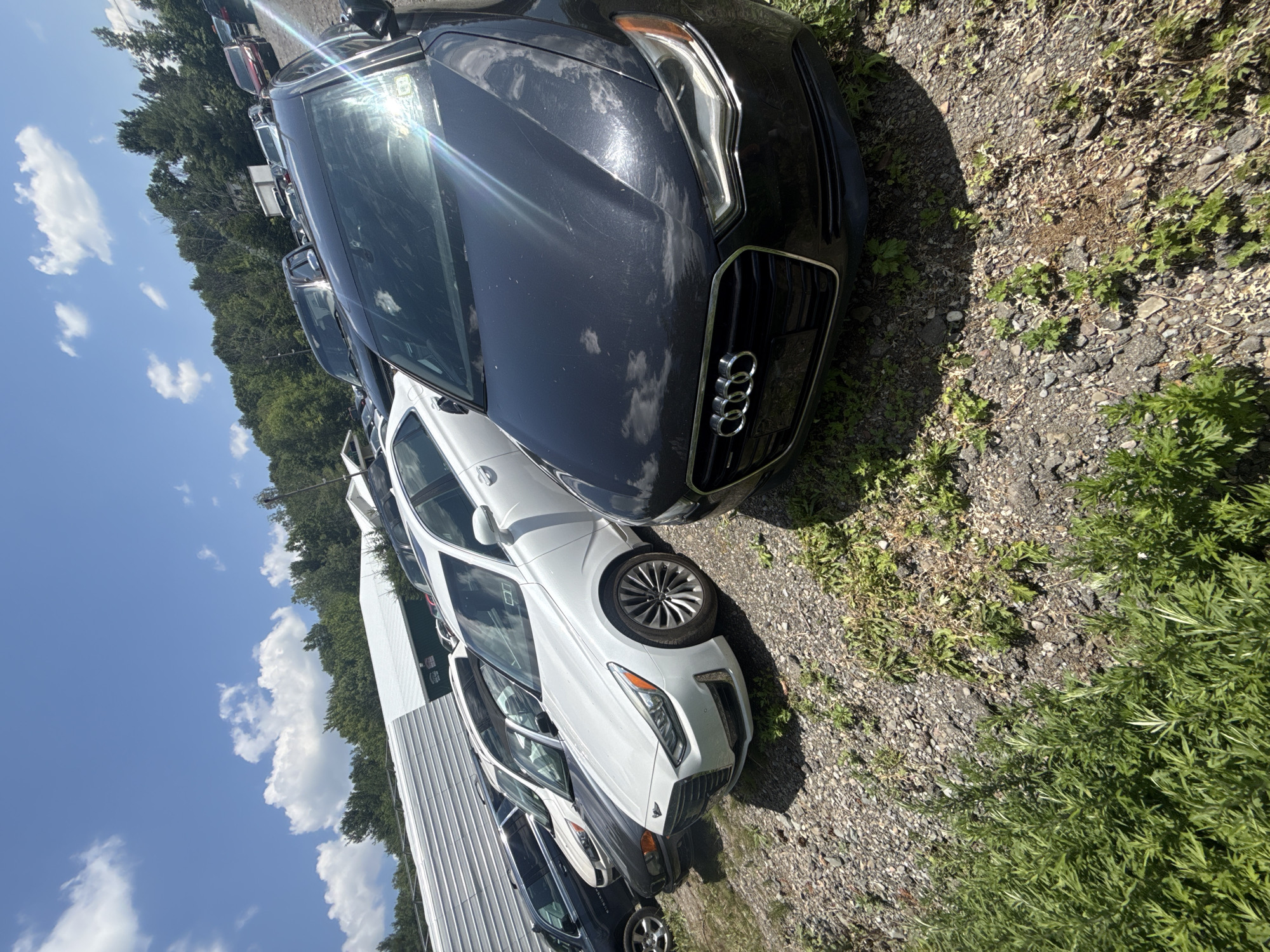 This is a ground-level, wide-angle shot of a car lot in the Village of Johnson City, United States of America, on a sunny day with scattered clouds. In the foreground, a dark grey Audi sedan is parked, facing towards the left of the frame. The car's grille, headlights, and hood are visible, reflecting the sky. Behind the Audi, several other vehicles are lined up, including a white sedan with dark accents on its hood, and further back, a dark SUV. In the background, a long, white, corrugated metal structure with a green section is visible, likely part of a building or storage area. A dense line of green trees forms a backdrop to the scene, with a clear blue sky overhead. The ground is covered in gravel and patches of green vegetation.