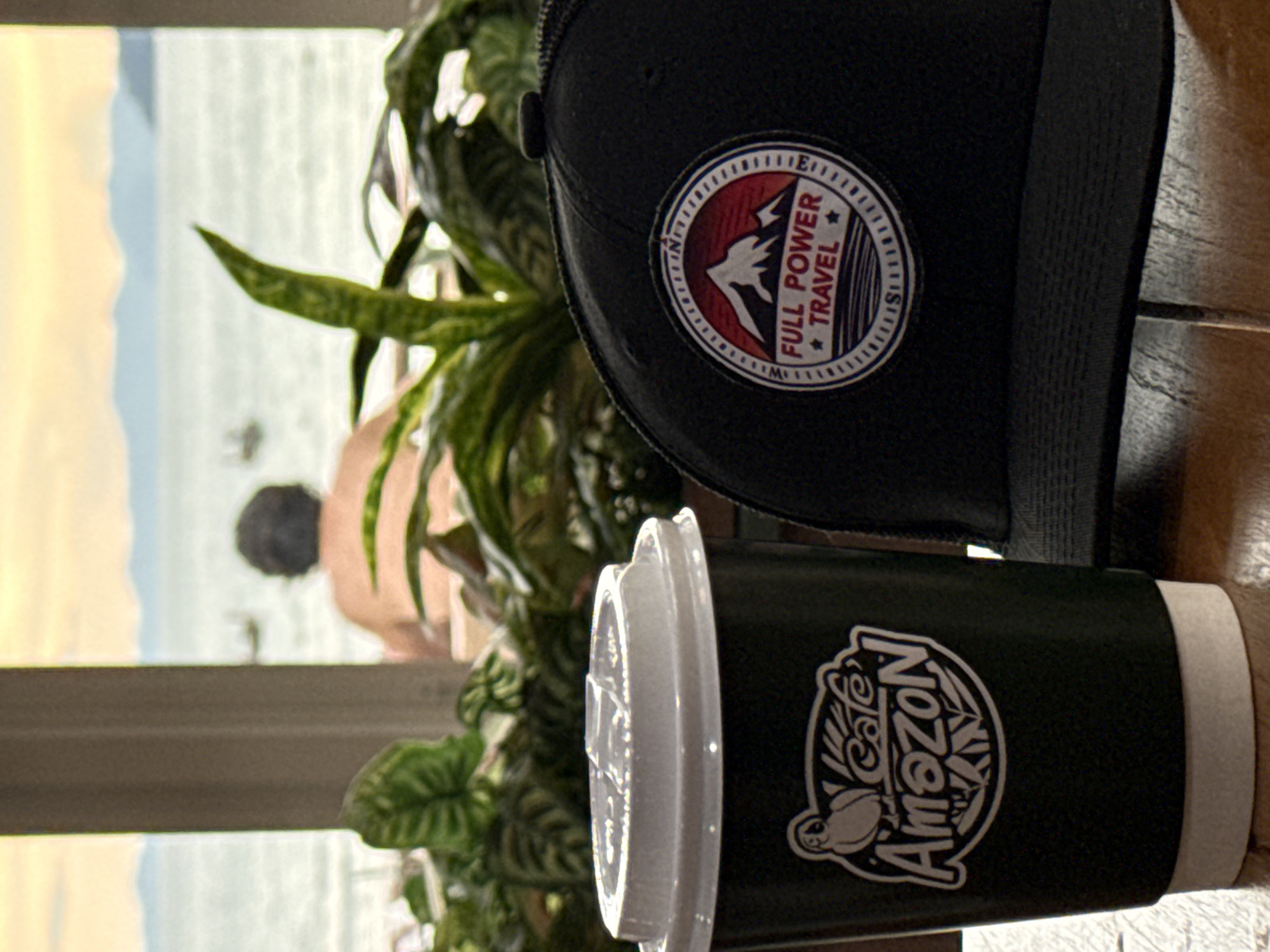 In the city of Ban Ao Nang, Thailand, a table in a waterfront cafe holds a dark green paper coffee cup with a white lid, featuring a logo with an owl and the text "Café Amazon." Beside the cup sits a black baseball cap with a circular patch that reads "FULL POWER TRAVEL" and depicts a mountain landscape. In the background, through a window, a person is visible from behind, seated and looking out at the sea. The sky is a soft orange and yellow, indicating either sunrise or sunset, and a hazy coastline with mountains is discernible in the distance. Lush green plants are positioned in front of the window, partially obscuring the view.