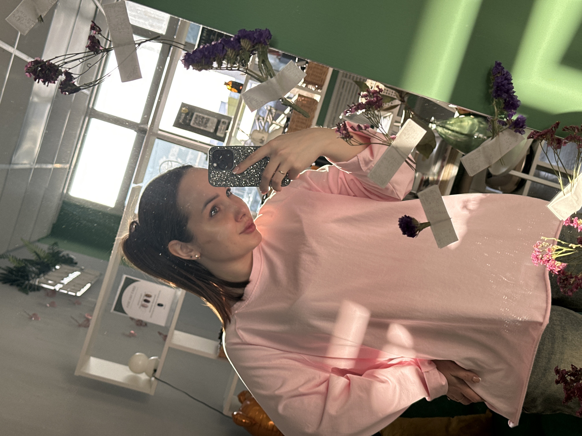 A female is depicted in a mirror selfie, standing indoors in what appears to be a beauty salon named "BCE SOCIETY" located at ul. Karla Marksa, 47, Chaikovsky, Perm Krai, Russia. She is approximately 20-30 years old, with dark brown hair styled in a high ponytail and wearing a loose-fitting, light pink long-sleeved shirt and blue jeans. Gold stud earrings are visible in her ears, and a gold-colored ring is on her left ring finger. She holds a dark-colored smartphone with a glittery case in her left hand, angled to take the photo, while her right hand rests on her hip. She is looking towards the phone with a subtle smile.
The mirror is large, rectangular, and features a green border on the right side. It is decorated with several sprigs of dried purple and pink flowers, secured with white adhesive tape. Reflected in the mirror is the interior of the establishment, revealing a light gray wall with a white open shelf unit on the left. On the shelf, a small white spherical lamp is visible, alongside a white rectangular sign with a green border and "BCE SOCIETY" text, displaying three circular icons and a vertical line with a dot. Above the shelf, a green leafy plant hangs. The reflection also shows a large window with multiple panes, through which bright daylight enters, and a white grid ceiling. Other reflections include wicker baskets, various signs (one with a QR code), and a brown bottle. The overall lighting is bright and natural, indicating daylight conditions.