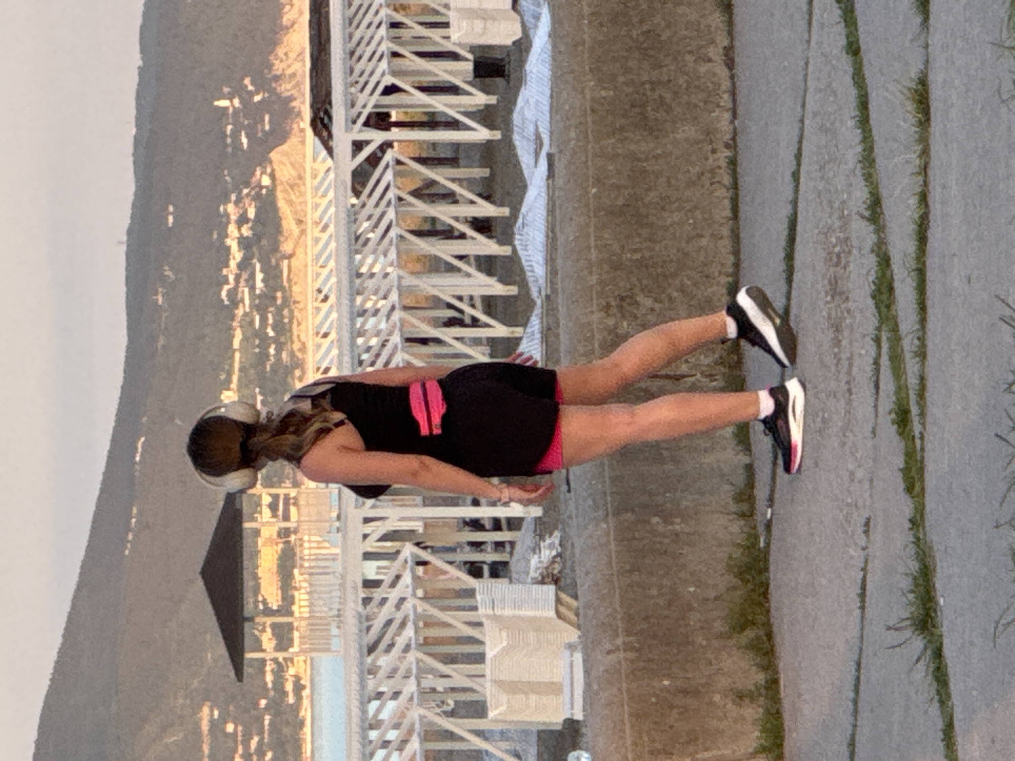 The image captures a woman standing on an outdoor paved pathway, overlooking a panoramic view of a city and mountains during what appears to be sunset or dusk. The woman is seen from a slight rear angle, wearing a black tank top, black shorts with pink trim, and running shoes with black, white, and pink details. She has a light-colored headband or headphones on her head.
To her right, a white wooden railing or fence extends across the scene, separating the pathway from the vista below. The background features a sprawling city with numerous buildings, illuminated by the warm, golden light of the setting sun, causing reflections and sparkles across the urban landscape. Beyond the city, large, dark mountains rise against the lighter sky. A small, dark, pyramid-shaped structure, possibly a gazebo roof, is visible among the city buildings.
The paved ground where the woman stands has distinct parallel lines, and patches of sparse green grass are visible along its edges. The scene suggests a scenic viewpoint or promenade in a city like Novorossiysk, Russia, known for its mountainous and coastal landscapes, at a tranquil time of day. There is no visible text in the image.