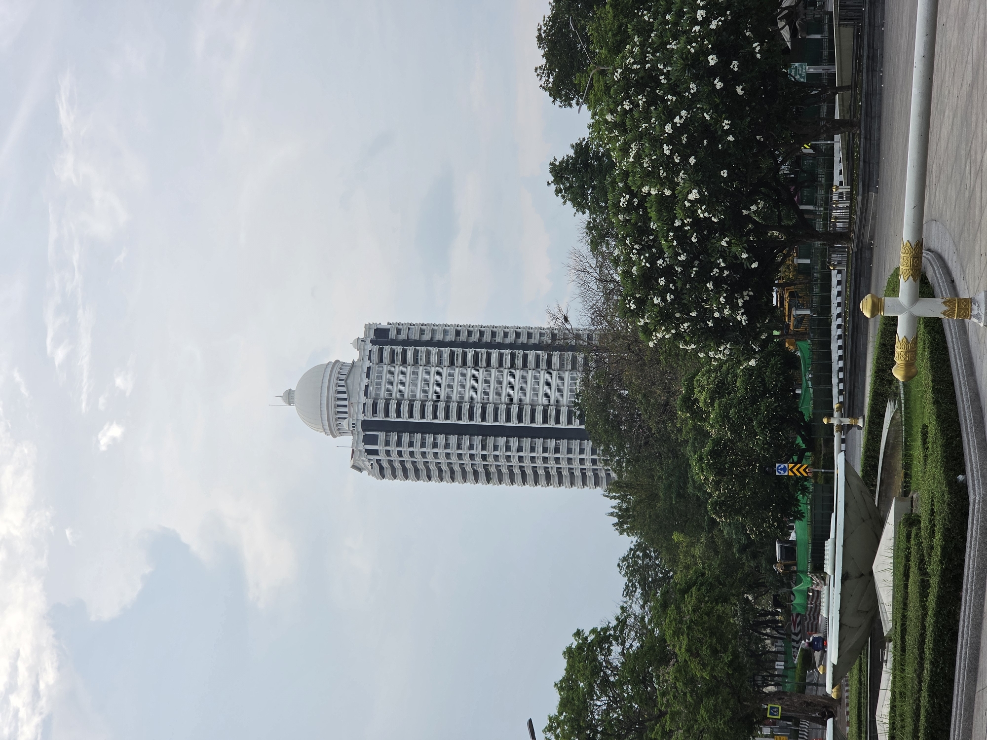A low-angle, outdoor shot, displays a tall white building in Bangkok, Thailand, interspersed with lush green trees on a partly cloudy day. The building, likely a high-end condominium or hotel, is vertically oriented, its white façade contrasting with black balconies and a distinctive domed top. The building’s base is partially obscured by dense trees.
On the right, more green trees in full foliage flank a street with a white cross-shaped barrier. The ground-level details are partially obstructed, but sections of shrubbery, sidewalks, and street are visible. A small sign and some construction netting are present behind the trees. The image is taken during daylight hours, with natural light highlighting the building's architectural details and the greenery.