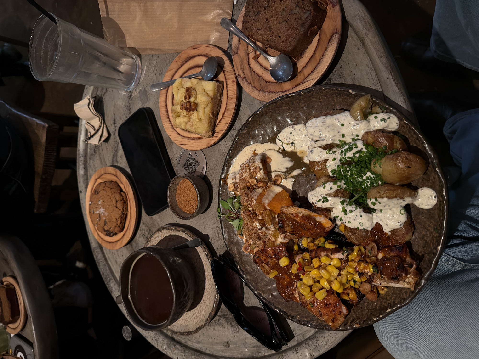 The scene captures a top-down view of a round, grey-toned table laden with food and beverages, likely within a restaurant or cafe setting. A large, dark-rimmed plate dominates the composition, containing a medley of roasted potatoes adorned with a creamy white sauce and fresh green chives, alongside what appears to be grilled chicken or other meat interspersed with corn.
To the left of the large plate, there is a cup of dark liquid, presumably coffee or tea, set on a small saucer. A small wooden plate holds a single cookie, while a cellular phone and a small bowl containing an orange granular substance (perhaps sugar or spice) rest nearby. Sunglasses are also laid on the table, adding to the sense of personal belongings amidst the food.
To the right, there's a wooden platter with a slice of what looks like apple pie, accompanied by a spoon. Next to it, another wooden platter holds a dark slice of bread. A tall glass stands empty on the left side of the table.
Based on the style of food, the rustic aesthetic of the table and servingware, and the overall ambiance, it's plausible the location is a trendy cafe or restaurant. However, without visual indicators of landmarks or specific decor, it is difficult to place this scene definitively within the city of London, United Kingdom.