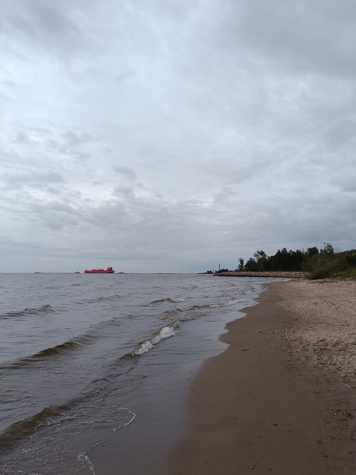 An overcast day scene at a sandy beach on the Baltic Sea, likely in Riga, Latvia. In the distance, a large, red cargo ship is visible on the water, accompanied by two smaller tugboats. To the right of the ship, a lighthouse and a treeline are discernible along the shore. The foreground features the gentle lapping of waves on the wet sand, with some dry sand further up the beach. The sky is filled with heavy, grey clouds. There are no people or animals visible in the image.