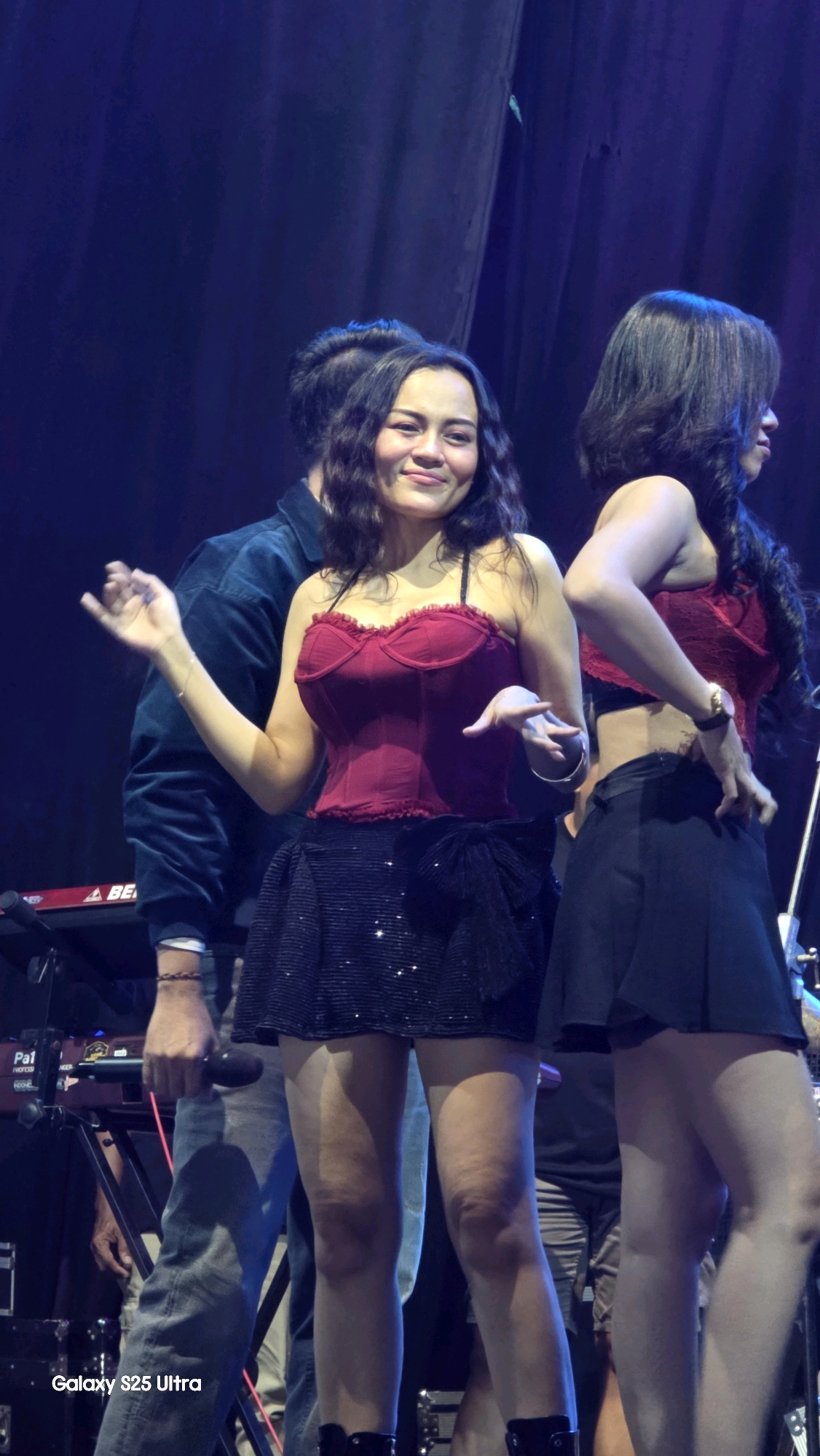 A group of people, possibly performers, are on a stage in what appears to be a concert venue. The focus is on two women in the foreground. The woman on the left is wearing a dark red corset-style top and a sparkly black mini skirt. She has dark, wavy hair and is smiling, with her right hand raised and her left hand gesturing. The woman on the right is wearing a similar dark red top and a black mini skirt, with her back partially to the camera. She has dark hair and is seen adjusting her skirt. Behind them, a person in a dark jacket is partially visible, holding a microphone. In the background, there are musical instruments, including a keyboard, and dark blue drapes. The lighting is dim and theatrical. The image displays a "Galaxy S25 Ultra" watermark in the bottom left corner. The location is Jaken, Indonesia.