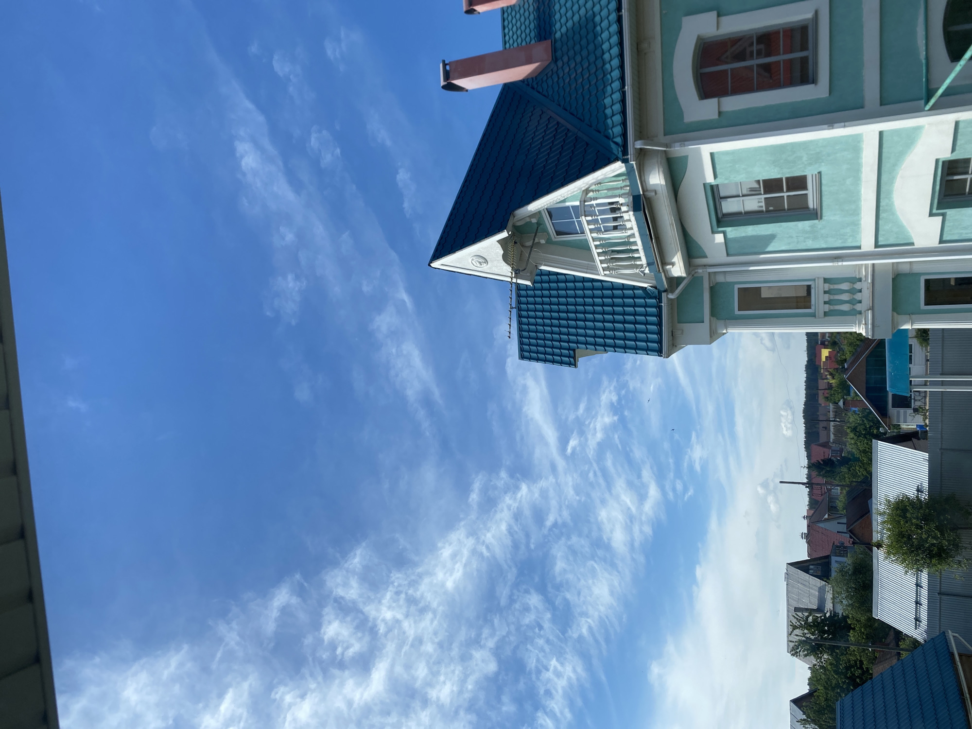 A bright daytime scene depicts a residential area, likely in a town or village. The most prominent feature is a light blue or mint green house with a dark blue tiled roof, occupying the right side and upper portion of the frame. This house exhibits decorative white trim, multiple windows including arched and rectangular shapes, and a small balcony with a white balustrade. A red chimney rises from its dark blue roof.
To the left, a vast expanse of clear blue sky with scattered wispy white clouds (cirrus type) dominates the view. In the mid-ground and background, a cluster of other houses with varying roof colors (red, grey) and some interspersed trees are visible, suggesting a populated area. Another dark blue tiled roof is partially seen in the lower left corner.
The lighting indicates it is daytime, probably mid-morning or afternoon, under clear and pleasant weather conditions. No specific activities or individuals are depicted, and there is no visible text. The overall impression is one of tranquility in a suburban or small-town environment, aligning with the provided location context of Chaikovsky, Russia.