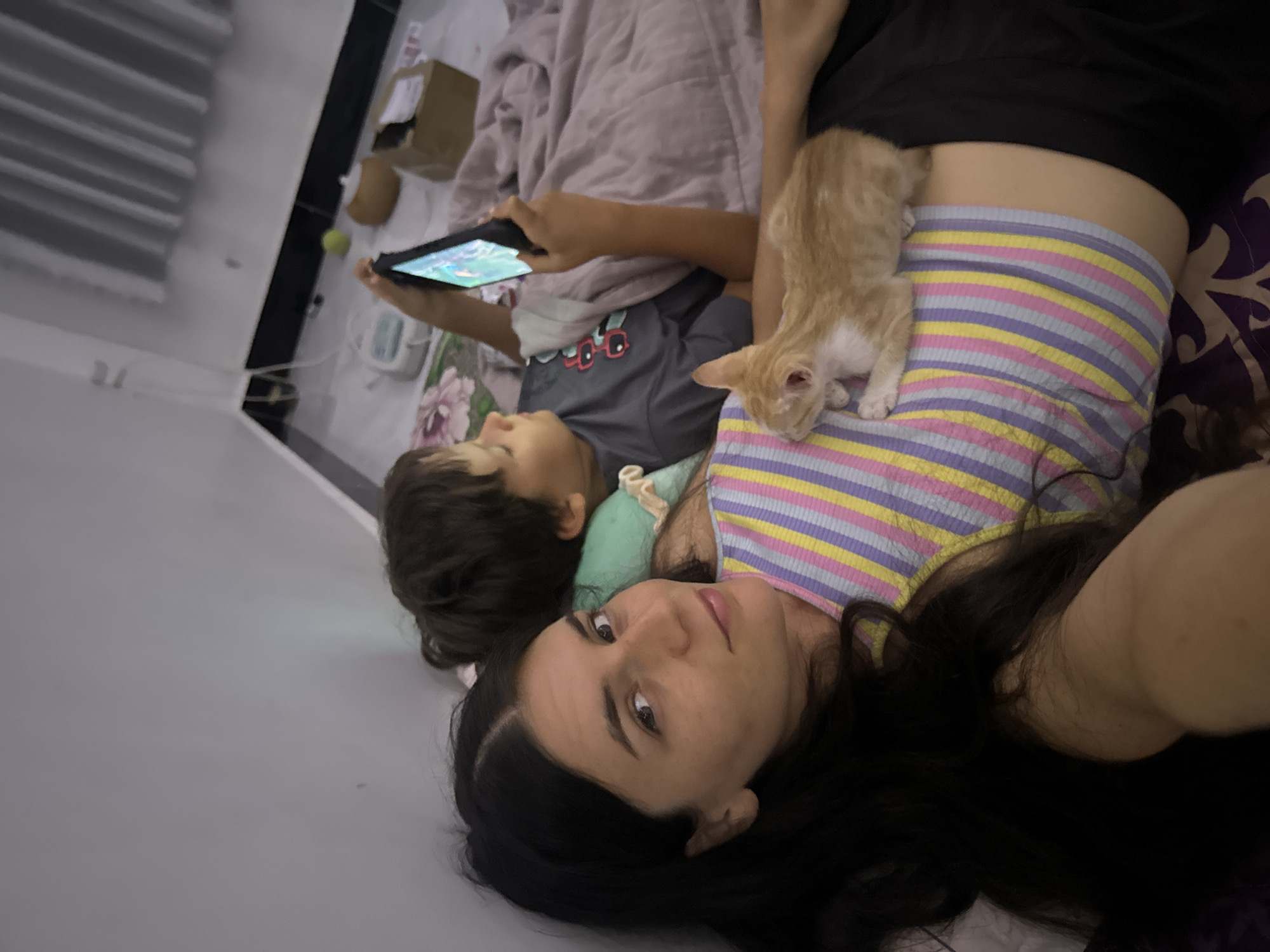 An overhead shot shows a woman and a child lying in bed in Mũi Né, Vietnam. The woman is on the left, her face turned towards the camera, and her long, dark hair is spread out. She is wearing a multicolored striped top. On her chest rests a small, ginger kitten. To the right of the woman and slightly behind her is a young boy, also lying in bed. He is looking down at a handheld gaming device he is holding, engrossed in playing. The background shows a bed with blankets and pillows. A wall is visible in the upper left corner, and a window with curtains is in the background to the right. Other items like a floor mat with a floral design, a yellow ball, and a cardboard box are scattered around the floor area. The overall scene depicts a relaxed domestic moment.