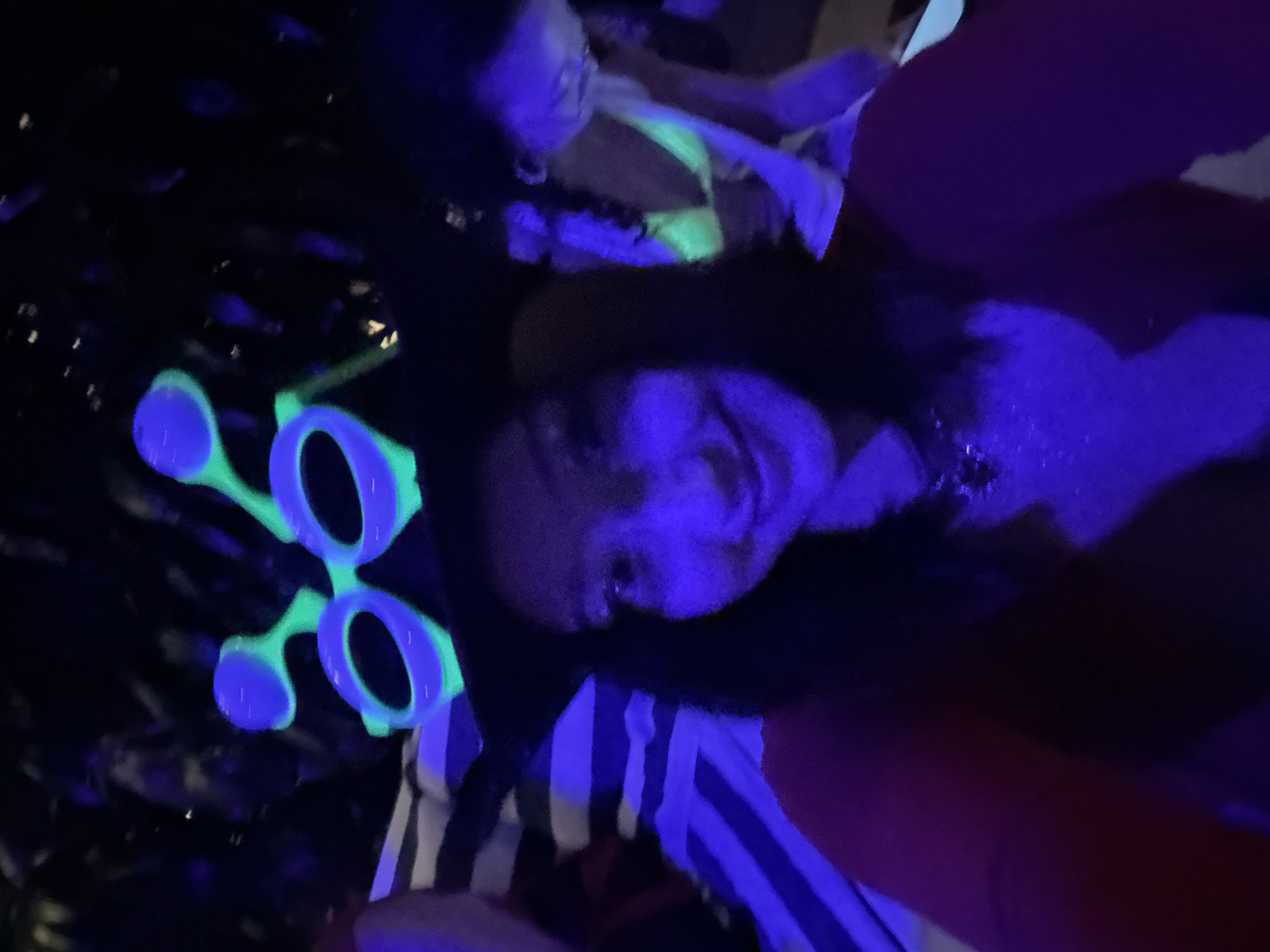 A selfie taken in a dark environment, illuminated with what appears to be UV lighting. The main subject appears to be a woman with dark hair, wearing a striped shirt. A pair of novelty glasses are also visible. Another person can be seen in the background. The location, Kuta Selatan, Indonesia is not determined by the image.