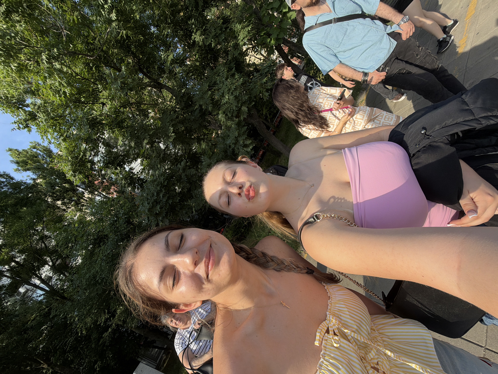 This media file features a vibrant outdoor scene on a sunny day, likely in Burgas, Bulgaria, dominated by a close-up selfie of two young women. The woman in the foreground, positioned lower in the frame, has brown hair and wears a yellow and white striped top, along with a delicate gold necklace featuring a small dagger pendant. Her eyes are closed, and she has a gentle smile, appearing relaxed. Behind her, a second woman with blonde hair is seen, wearing a purple strapless top. Her eyes are also closed, and her lips are pursed in a playful manner, as if blowing a kiss. Both women have tan skin, indicating they are enjoying the warm weather.
In the mid-ground and background, several other individuals are visible. These include a man in a light blue shirt, black pants, and a dark cap, and a woman in a light-colored patterned dress. Further back, another person is wearing a dark top with some indistinct white text. A black puffer jacket and a black shoulder bag with a gold chain strap are also visible in the foreground, belonging to the women.
The setting is characterized by abundant green trees with dense foliage, suggesting a park or a tree-lined street. A clear blue sky is visible through the tree canopy. Paved ground, consistent with a sidewalk or plaza, can be seen, with shadows indicating bright sunlight, likely midday or early afternoon. The overall mood is casual, relaxed, and summery. No clearly legible text is prominently visible in the image.