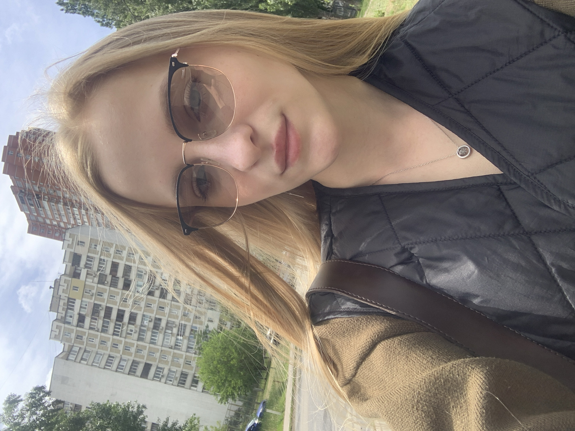 A selfie taken in Kyiv, Ukraine, features a young woman with long blonde hair wearing glasses and a black quilted vest. She has fair skin and is wearing a silver necklace with a circular pendant. Part of a brown purse strap is visible. In the background, multi-story apartment buildings are seen, as well as green trees and a patch of sky. The camera is tilted.