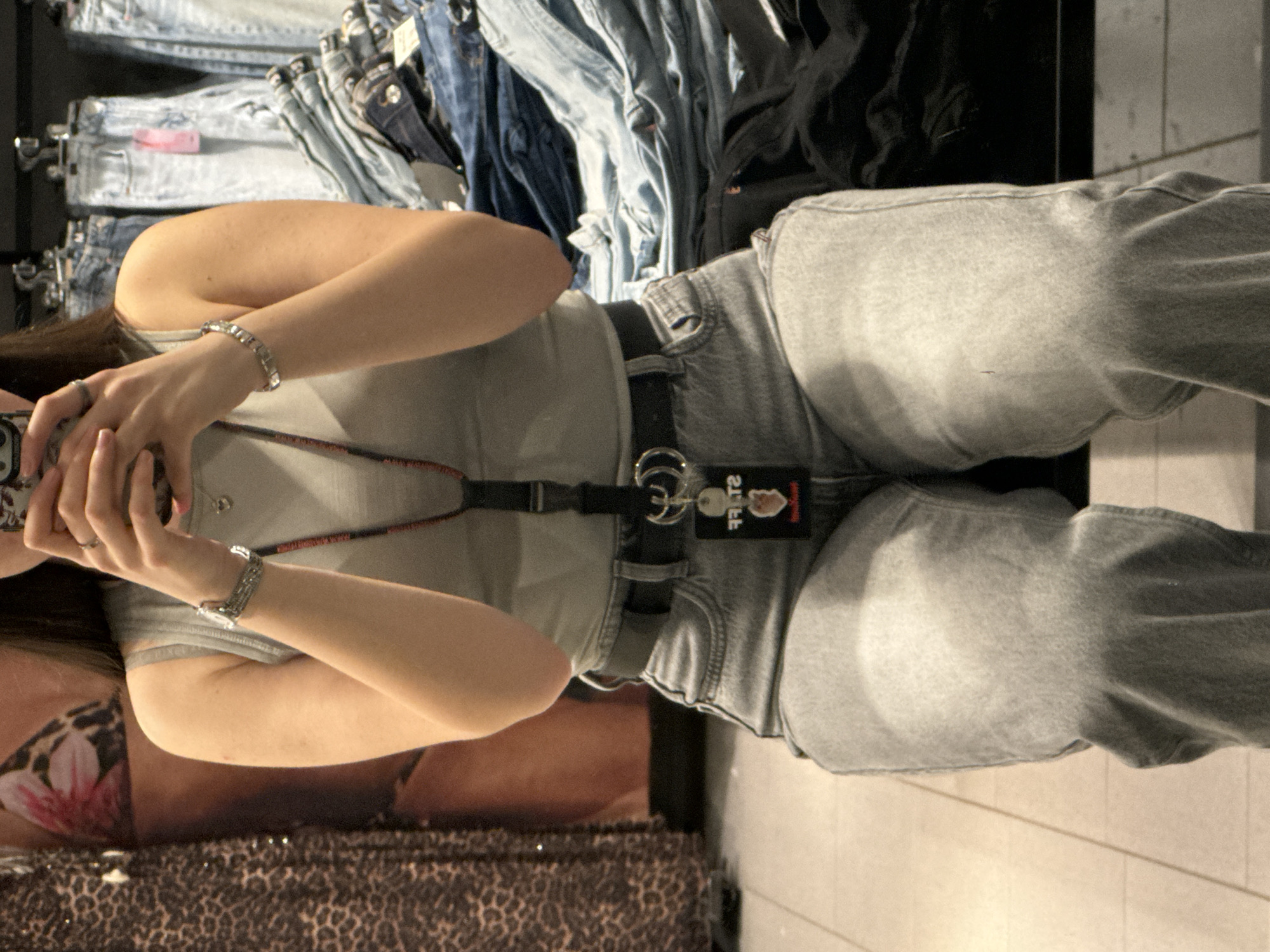 A person wearing light-colored jeans, a gray t-shirt, and a black belt stands in front of a clothing rack filled with denim jeans. They are holding a mobile phone, presumably taking a picture in an indoor setting. Part of a display with a leopard print and a pink flower is visible at the bottom left. The location is not specified.