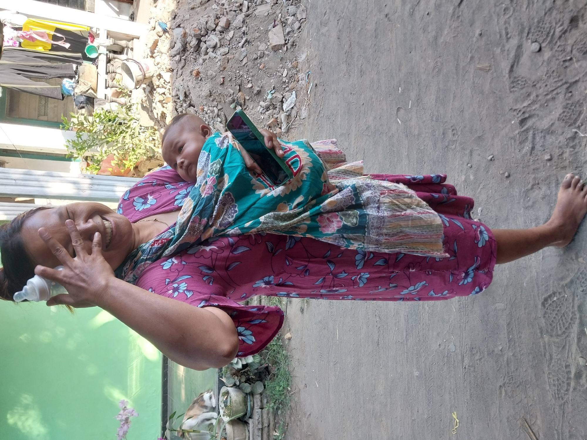 A woman in Nglegok, Indonesia, is holding a baby in a sling while standing on a dirt path. The woman is wearing a dark pink floral dress and has her hand over her face, holding a baby bottle. The baby is looking up. Behind them, there is a green wall, some plants in pots, and a pile of rubble. The scene suggests a casual, everyday moment in a residential area.