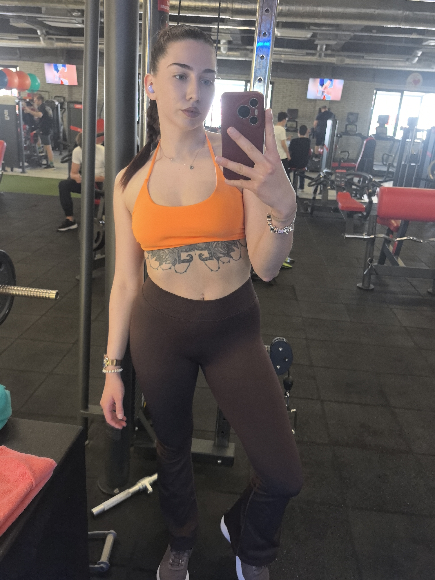 A woman with dark hair pulled back in a braid and wearing an orange halter top and brown leggings takes a selfie in what appears to be a gym. She is holding a phone in her right hand. The gym has typical exercise equipment, including weight machines and treadmills. Other people are visible in the background. The exact location within Stara Zagora, Bulgaria, is not discernible from the image.