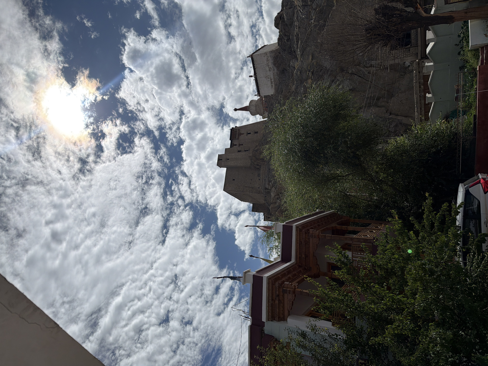 The image shows a view of the Tsemo Monastery in Leh, India. The monastery is situated on a hill, with a large fortress-like structure at the top. In the foreground, there is a traditional Ladakhi building with intricate wooden carvings. The sky is cloudy with the sun shining through, creating a dramatic effect. A white car is parked in front of the building. The overall scene depicts a blend of architecture and natural landscape, characteristic of Leh.