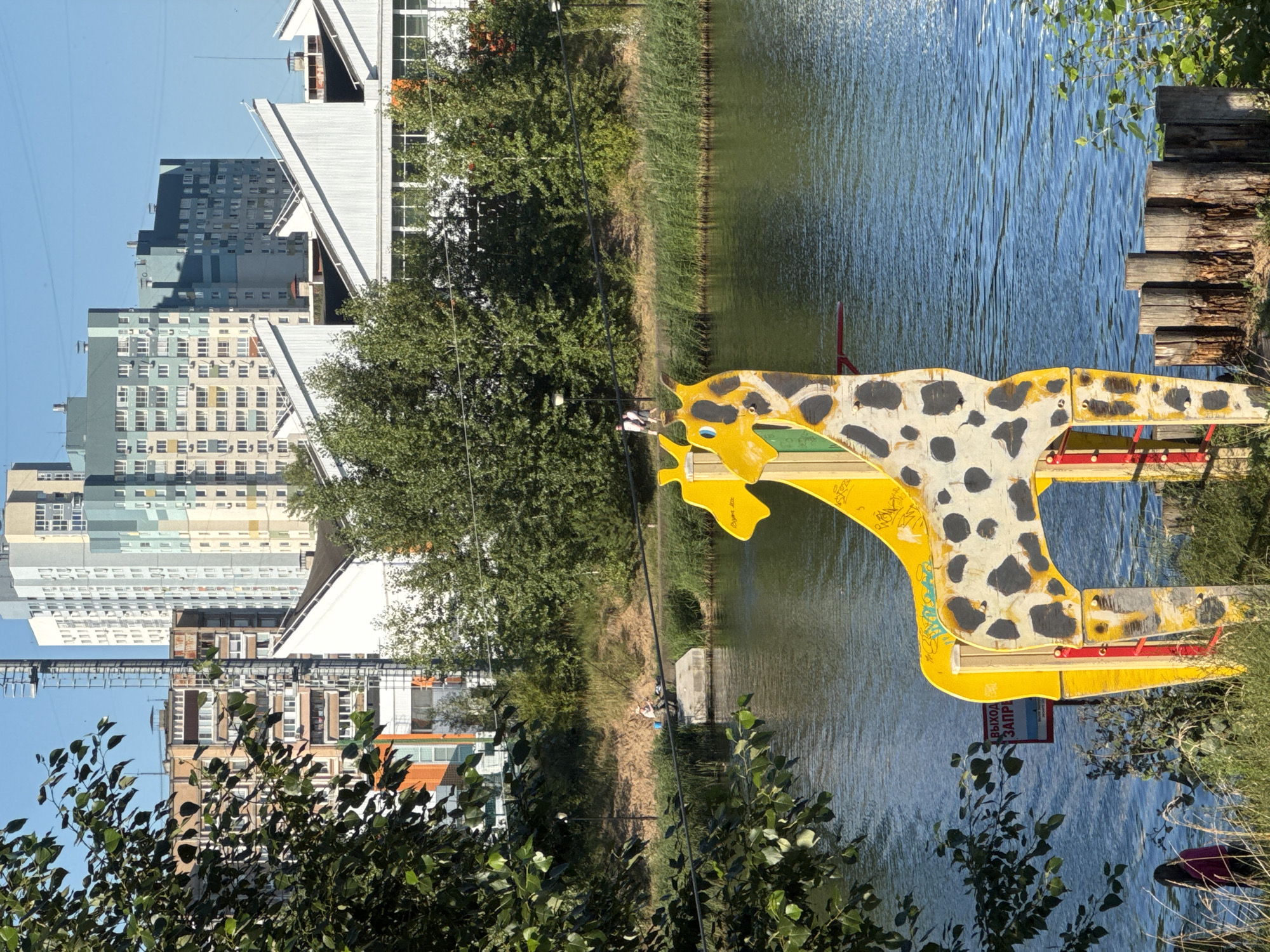 The image depicts a waterside scene with a large, yellow, giraffe-shaped playground structure in the foreground. The structure is partially painted with black spots and shows signs of wear and tear, with visible graffiti. In the background, a body of water, likely a river or canal, is visible, with a grassy bank and trees lining it. Further in the background, a cluster of modern, high-rise buildings, typical of urban development, are silhouetted against a clear blue sky. A man is seen fishing from a concrete platform on the bank. The overall setting suggests a recreational area within a city, likely Nizhny Novgorod, Russia, given the architectural style of the buildings and the presence of a body of water within an urban environment.