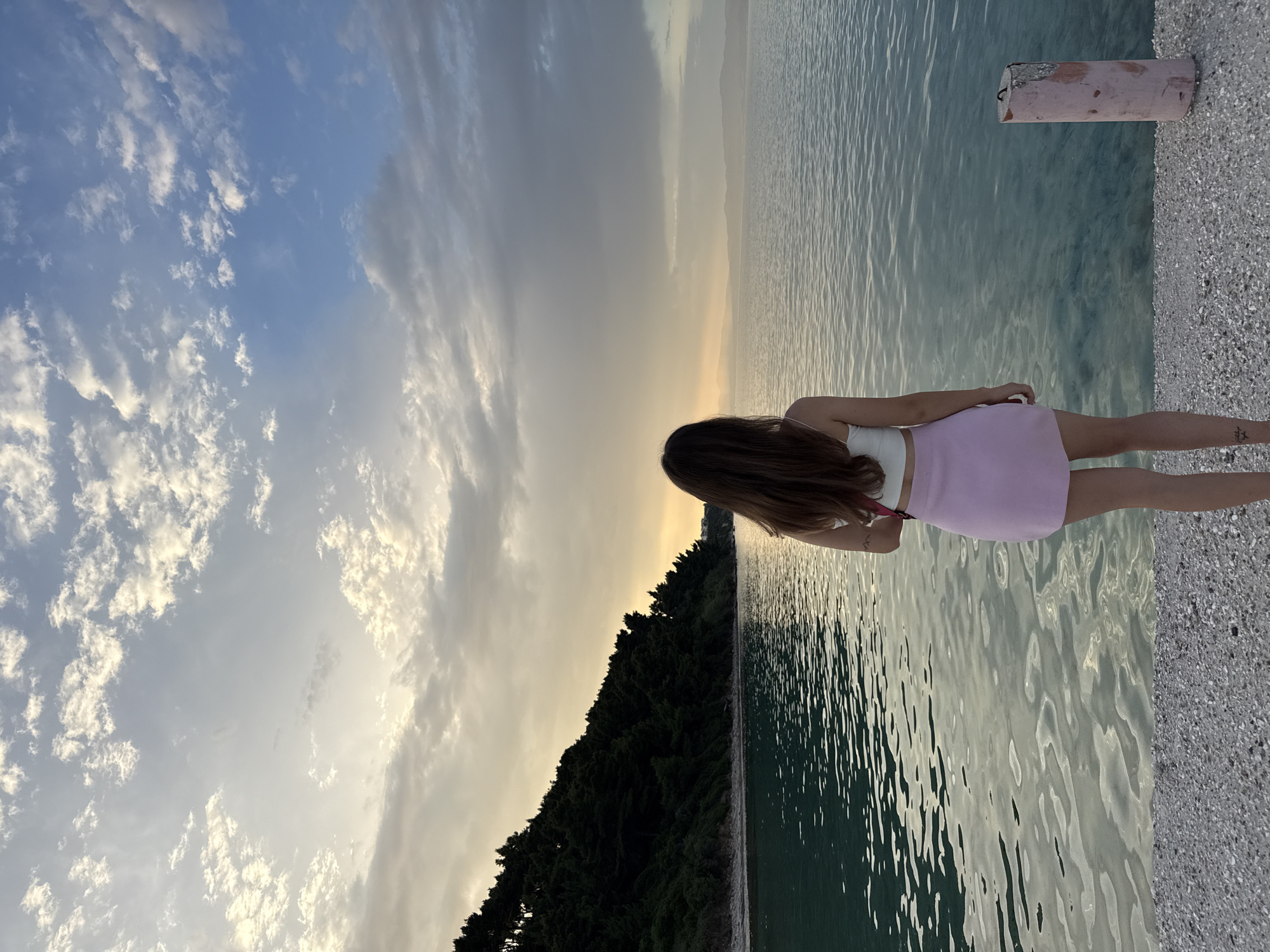 A woman stands on a pebble beach, her back to the camera, facing a calm, turquoise sea. She has long, brown hair and is wearing a white crop top and a light pink skirt. The sea reflects the soft light of a cloudy sunset, with the sky displaying hues of yellow, orange, and blue. To the left, a densely forested hill slopes down towards the water. In the distance, hazy mountains are visible on the horizon. On the right side of the frame, a weathered, pink-painted bollard stands on the edge of the beach. The scene appears to be a tranquil coastal area in Kassandra Municipal Unit, Greece, likely during the evening.