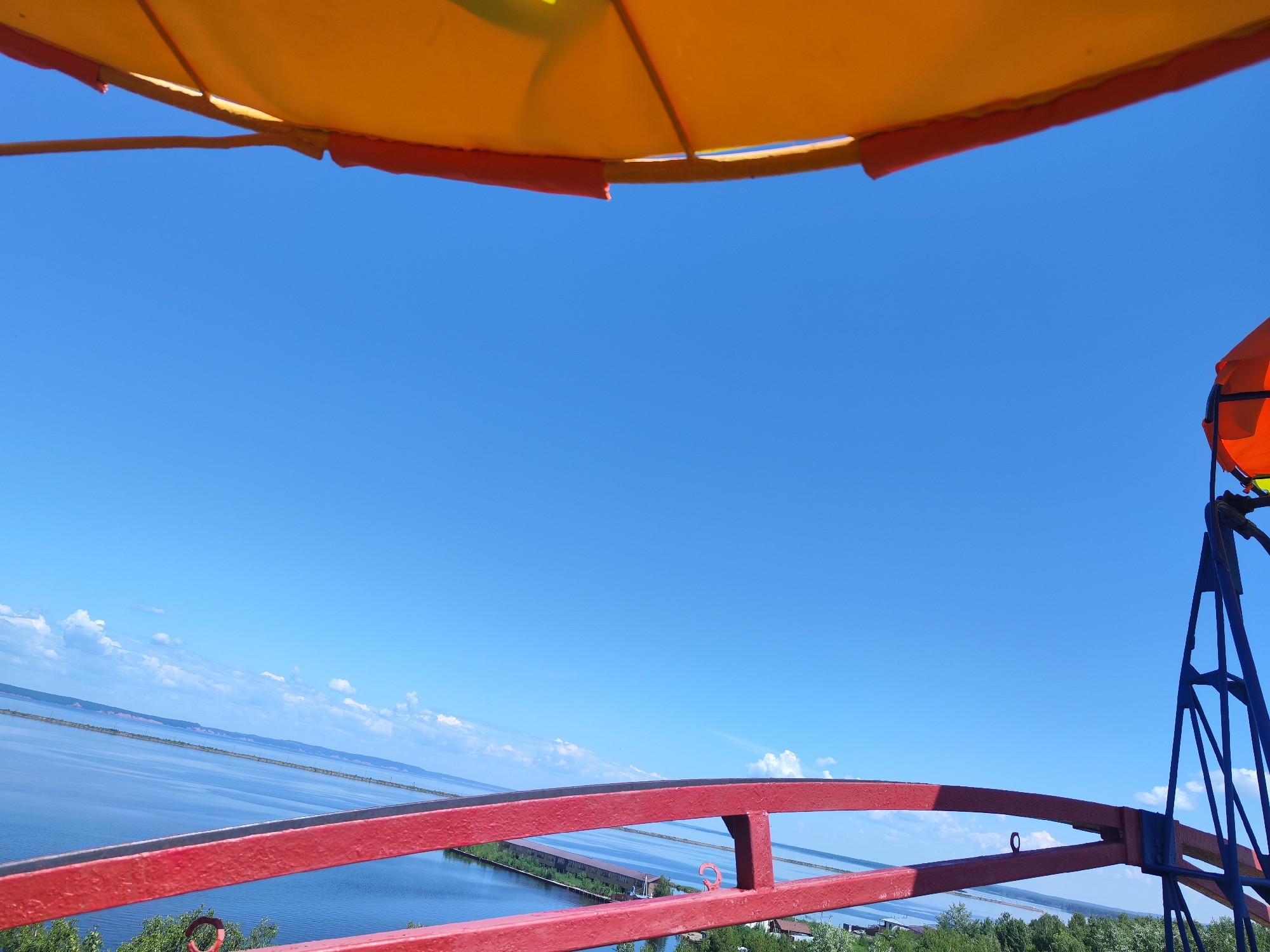 This is an outdoor, eye-level, medium shot of a view from a tall structure, possibly a Ferris wheel, in Chaikovsky, Russia. In the foreground, a bright red railing with some structural supports is visible. Above this railing, a large expanse of blue sky dominates the upper half of the image. At the very top, a yellow and orange canopy is partially visible, suggesting it is part of the structure the camera is on. To the right, a blue metal support structure and an orange object are visible, likely part of the same attraction. In the middle ground and background, a wide body of water, possibly a river or reservoir, stretches horizontally across the frame. The water is calm, reflecting the blue sky and some white clouds. On the far side of the water, a thin strip of land with some vegetation and possibly buildings is visible, with a distant hazy horizon. The overall impression is a sunny day with a panoramic view.