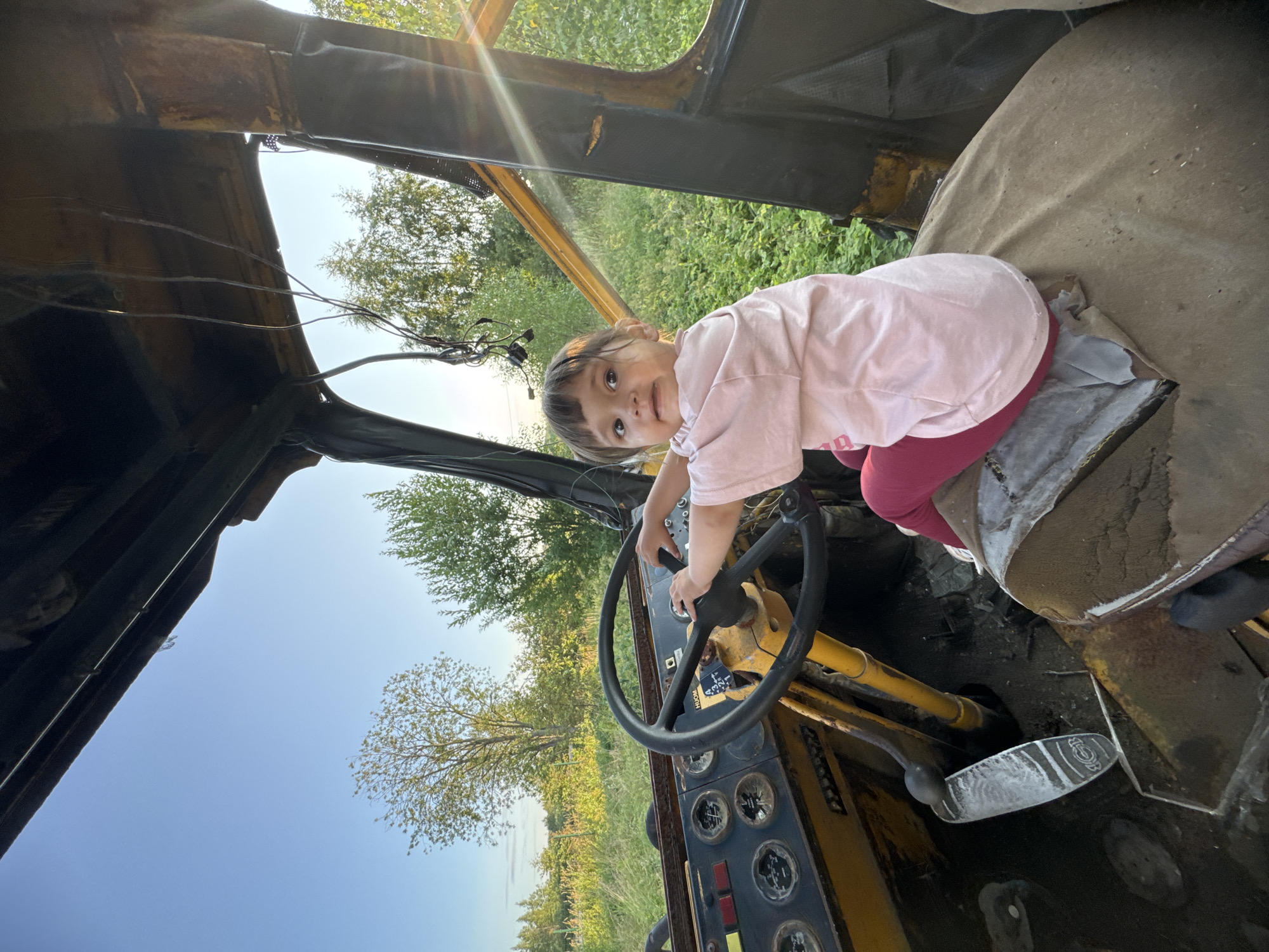 A young child is seated in the driver's seat of an old, weathered tractor, possibly in a rural or outdoor setting. The child, wearing a pink t-shirt and maroon leggings, has their hands on the steering wheel, appearing to be playing or pretending to drive. The tractor's interior is worn and rusty, with visible gauges and pedals. The background shows a natural landscape with trees and vegetation under a clear blue sky. The image is taken outdoors in Innolovo, Russia.