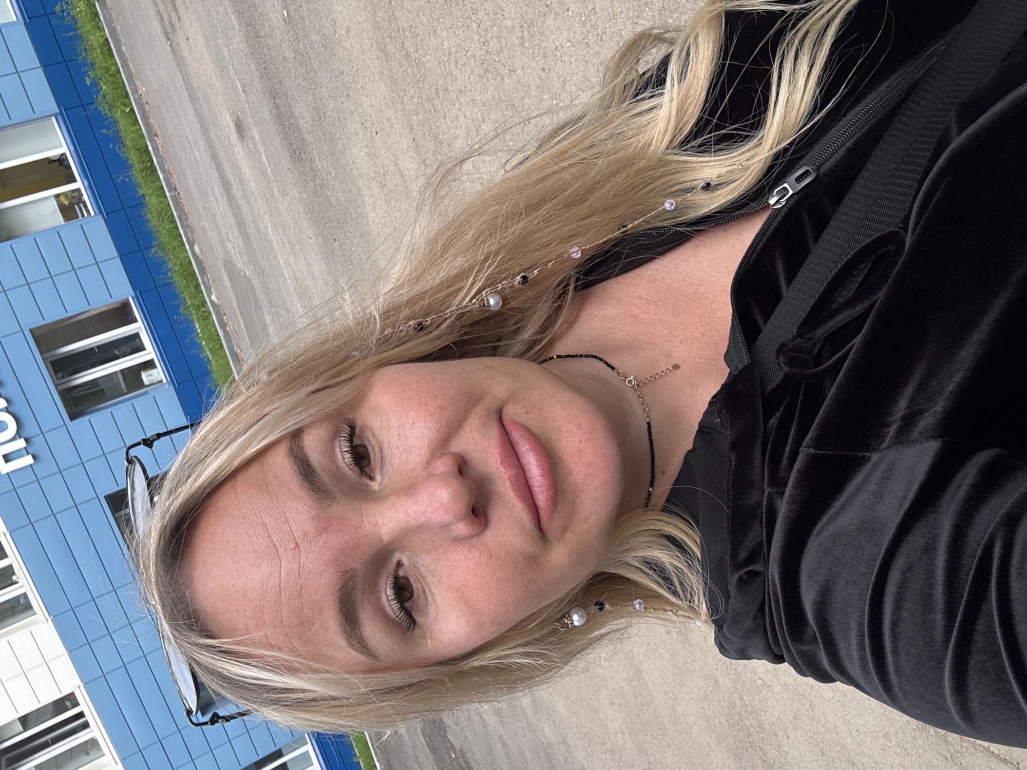 A young adult Caucasian woman with blonde hair, wearing a black velour tracksuit and sunglasses, takes a selfie outdoors. She is standing in front of a blue and white building with windows, likely in the city of Chastyye, Russia. The ground is paved with asphalt. Her hair is decorated with beads and pearls, and she is wearing a black choker necklace with a pendant. The overall scene suggests a casual, daytime setting.