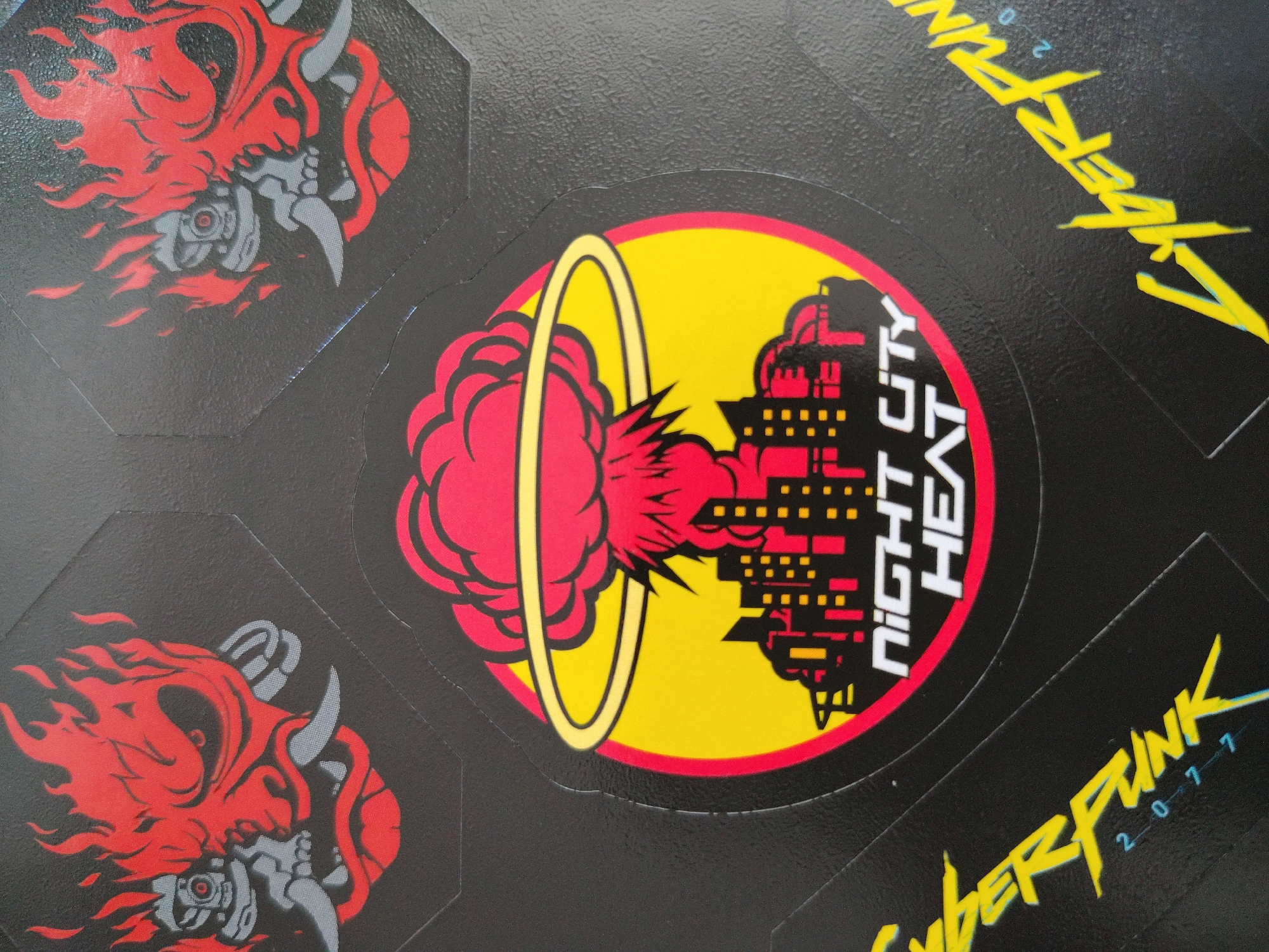The image displays a close-up view of several stickers, likely part of a collection or a product packaging. The stickers are predominantly black with vibrant graphic designs.
One prominent sticker features a circular emblem with a yellow outer ring and a bright yellow background. Inside this ring, a red, cloud-like explosion is depicted, with a stylized yellow ring encircling the top portion of the explosion. To the right of the explosion, a cityscape is illustrated in red and yellow, with numerous rectangular windows suggested by yellow dots and lines. The text "NIGHT CITY HEAT" is printed vertically in white on the cityscape.
Another set of stickers showcases a recurring motif of a demonic or monstrous head with flowing red flames emanating from it. The head is rendered in red and gray, with sharp horns and glowing red eyes. The texture of the stickers appears to be somewhat glossy, with a subtle embossed pattern on the black background.
The overall aesthetic of the stickers suggests a theme related to a video game or a similar form of media, possibly "Cyberpunk 2077" given the visible text "CYBERPUNK 2 0 7 7" in a distinctive yellow font along the bottom right of the image. The colors are bold and high-contrast, with a clear emphasis on red, yellow, and black. The image does not provide information about the time of day, weather, or a specific emotional tone beyond the aggressive imagery. The location context of Kolomyia, Ukraine is external information and not derivable from the image itself.
