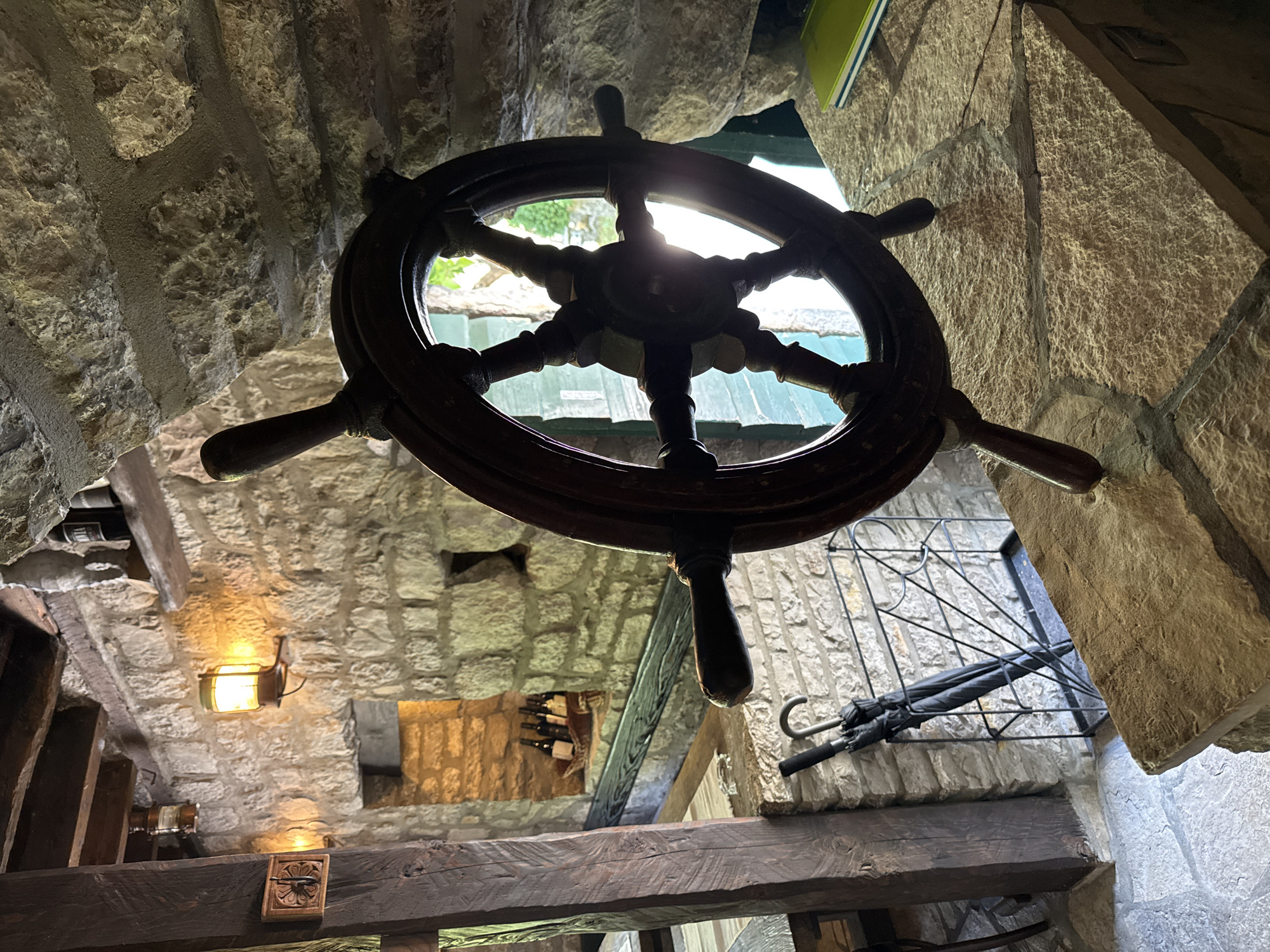 The image depicts the interior of a rustic stone building, likely a restaurant or bar, located in Morinj, Montenegro. A large, dark wooden ship's wheel is the central focus, positioned on a stone ledge. To the left of the wheel, an umbrella stand holds two closed black umbrellas. Behind the umbrellas, a stone wall is visible with a metal grate. Further to the left, a heavy wooden beam extends upwards, and a small decorative wooden carving is attached to it. An illuminated lantern hangs on the stone wall. In the background, a recessed shelf holds several bottles of wine in a basket. Sunlight filters through a window or opening behind the ship's wheel, creating a lens flare effect. The overall atmosphere is dimly lit and aged, suggesting a historical or nautical theme.