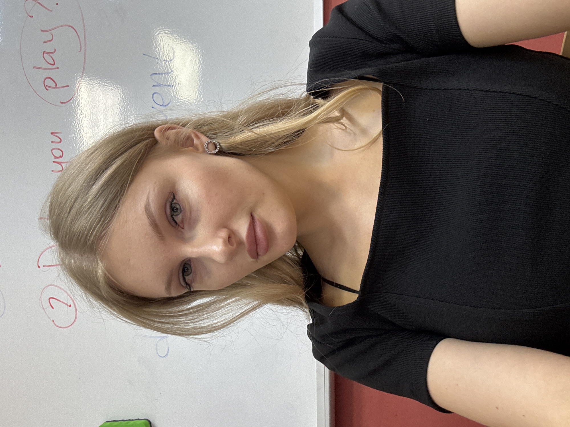 A blonde woman wearing a black dress stands in front of a white board. The whiteboard has writing on it in red and blue. The writing includes the words "you" and "(play)?". The background includes a red wall. The image may have been taken in the city of Perm, Russia, but there are no indicators in the image itself.