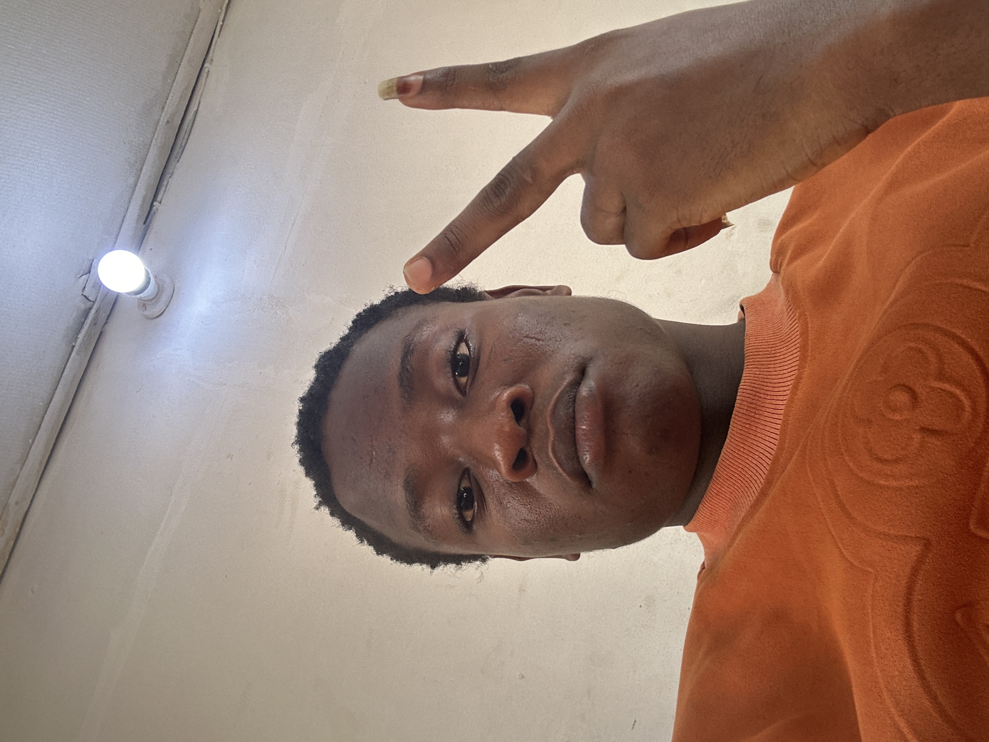 A low-angle selfie of a young Black man with short hair and an orange sweater making a peace sign on his head. He is indoors, with a light fixture visible on the ceiling above. The location is unknown.