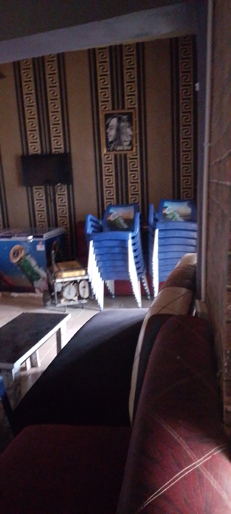 The media file shows an indoor scene, likely a lounge or waiting area. In the foreground, a dark red and black upholstered chair is visible, suggesting a comfortable seating arrangement. Behind it, a black table with white legs stands prominently. Further back, several stacks of bright blue plastic chairs are arranged, some with what appear to be images on the backrests. To the left, a blue refrigerator with a pipe or hose attached is partially visible, and next to it is a yellow piece of machinery. The walls are covered in striped wallpaper with a decorative border. A framed picture, possibly a portrait, hangs on the wall above the wallpaper. A dark rectangle on the wall to the left of the picture may be a television. The lighting appears dim, suggesting it could be evening or an indoor environment with controlled lighting. There is no indication of people or specific weather conditions. No visible text is discernible.
