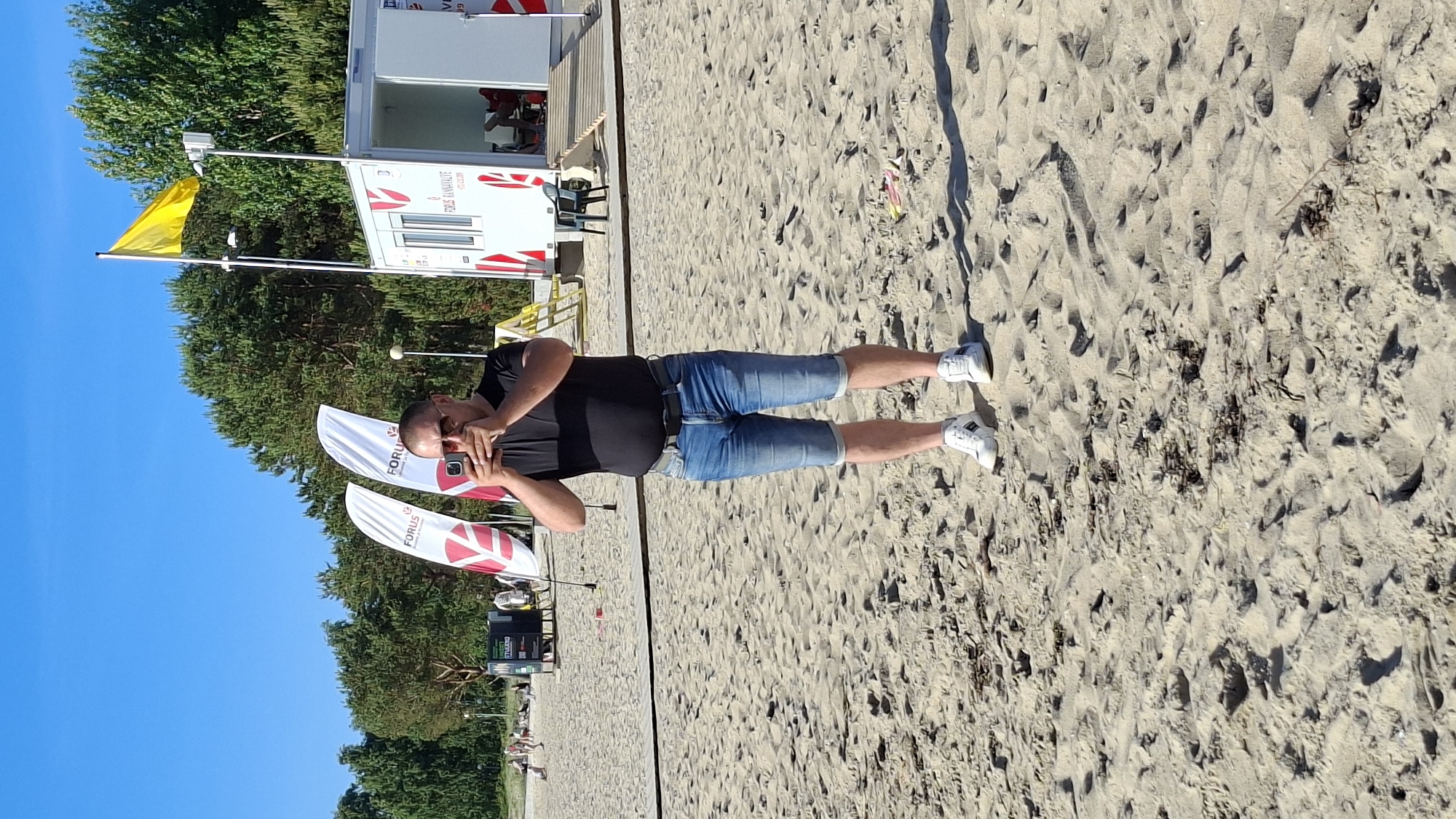 A man stands on a sandy beach in Tallinn, Estonia, holding a mobile phone to take a photo. He is wearing a black t-shirt, blue denim shorts, a belt, and white sneakers. Behind him, there are several white flags with red logos, a white portable building, and a yellow flag on a pole. The sky is clear blue, and trees are visible in the background. The beach is covered in sand with some dry seaweed and debris.