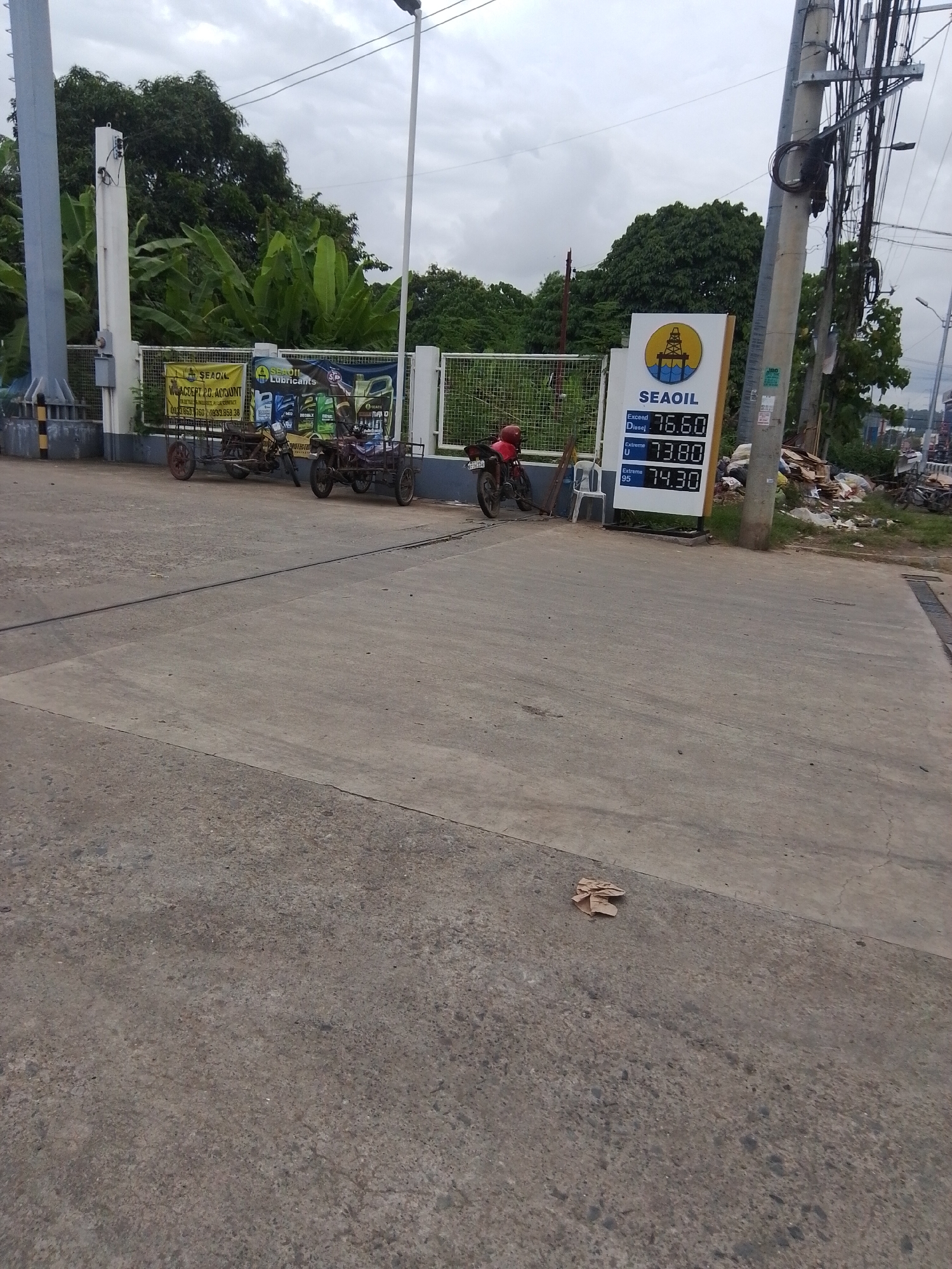 The image shows a SeaOil gas station in Davao City, Philippines. In the foreground, a concrete pavement stretches across the scene. In the background, a white gas price display board shows prices for different types of fuel. A red motorcycle with a license plate is parked to the left of the board, and a white plastic chair is placed next to it. Further to the left, two tricycles are parked. Behind them, a fence and lush green trees can be seen. Electrical poles and wires are visible on the right side of the image, along with some discarded debris. The sky is overcast.
