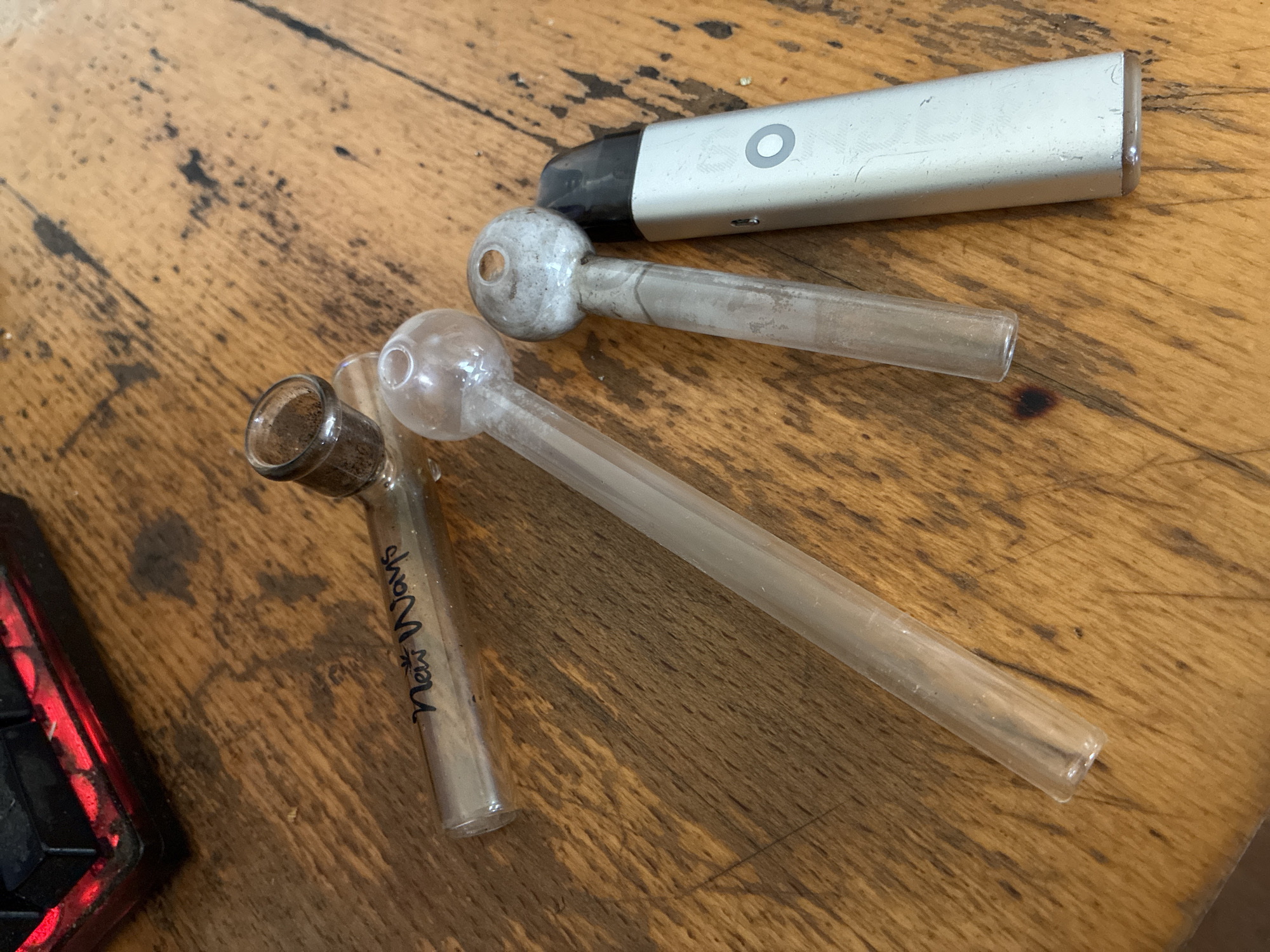 The image displays several items laid out on a weathered wooden surface, likely a table or floor, characterized by visible grain and dark stains. There are no people visible.
The primary subjects are three glass pipes and one electronic vape pen. The glass pipes are transparent with a bulbous end featuring a small hole, connected to a straight stem. Two of the pipes show significant internal residue, appearing white or greyish, consistent with byproducts from smoking or vaporizing. One of these two pipes also has a darker, burnt residue within its bulb. The third glass pipe is smaller, has a more uniform tubular shape with a slightly wider opening at one end, and contains dark, granular residue. On this smaller pipe, the text "New Ways" is handwritten in black marker.
The vape pen is silver-colored with a black mouthpiece and cap, showing signs of wear such as scratches. It has a circular button or indicator light on its body.
Part of a red object, possibly an electronic device like a keyboard or gaming console, is visible in the lower left corner, featuring red backlighting or trim.
There is no activity taking place; the items are static. The lighting suggests an indoor setting. No specific time of day or weather conditions can be inferred. While the location context is given as Varna, Bulgaria, the image itself does not contain any visual cues to confirm this geographical location.