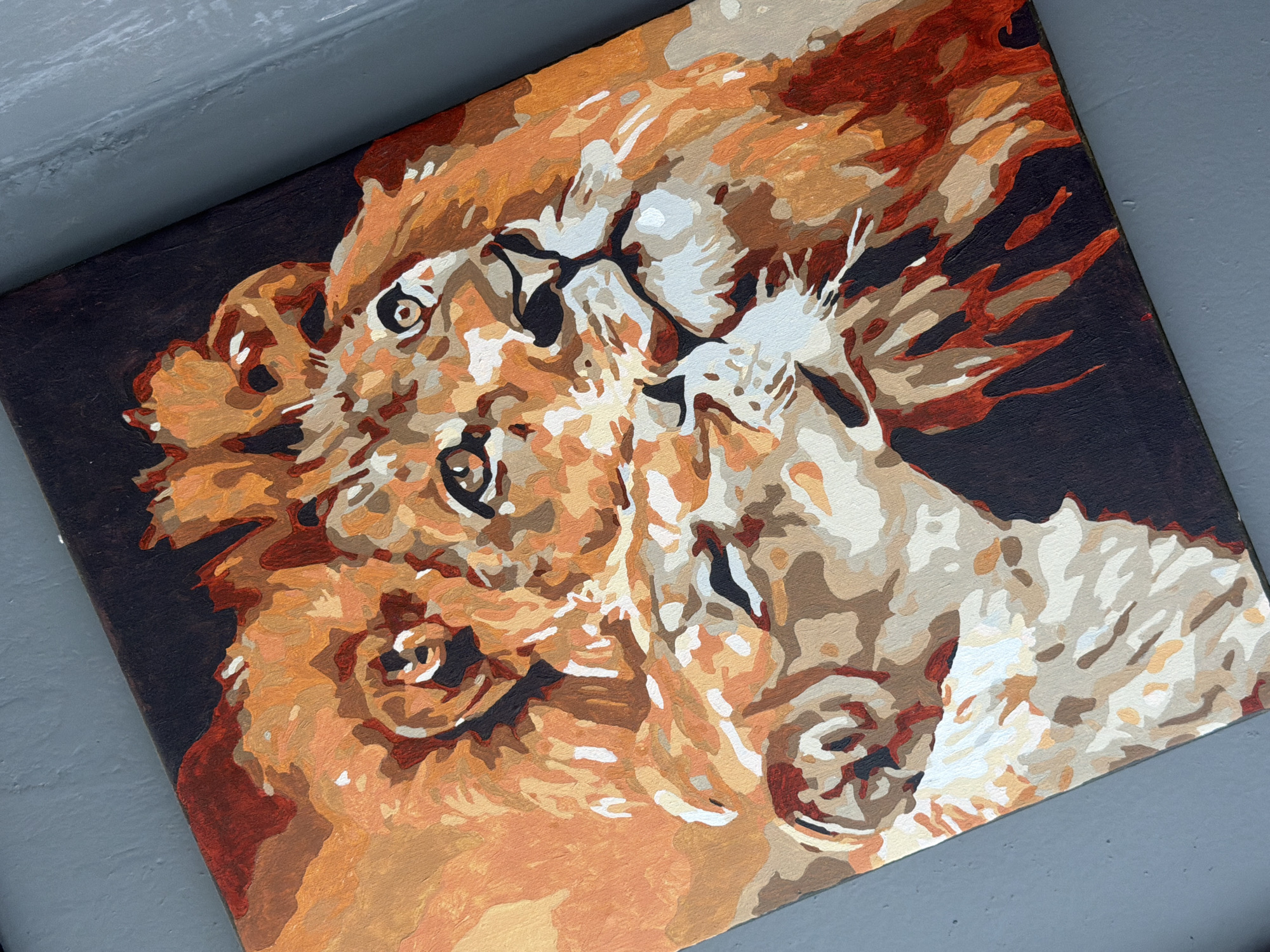 The image is an amateur painting of lions, rendered in a paint-by-numbers style, lying on a gray surface. The painting depicts a pair of lions with stylized manes, featuring various shades of brown, tan, and cream against a dark brown background. The composition is dense, with the lions' forms overlapping. The painting appears to be resting on a gray surface, possibly concrete or metal. There is no indication of the city of Rostov-on-Don, Russia in the image.