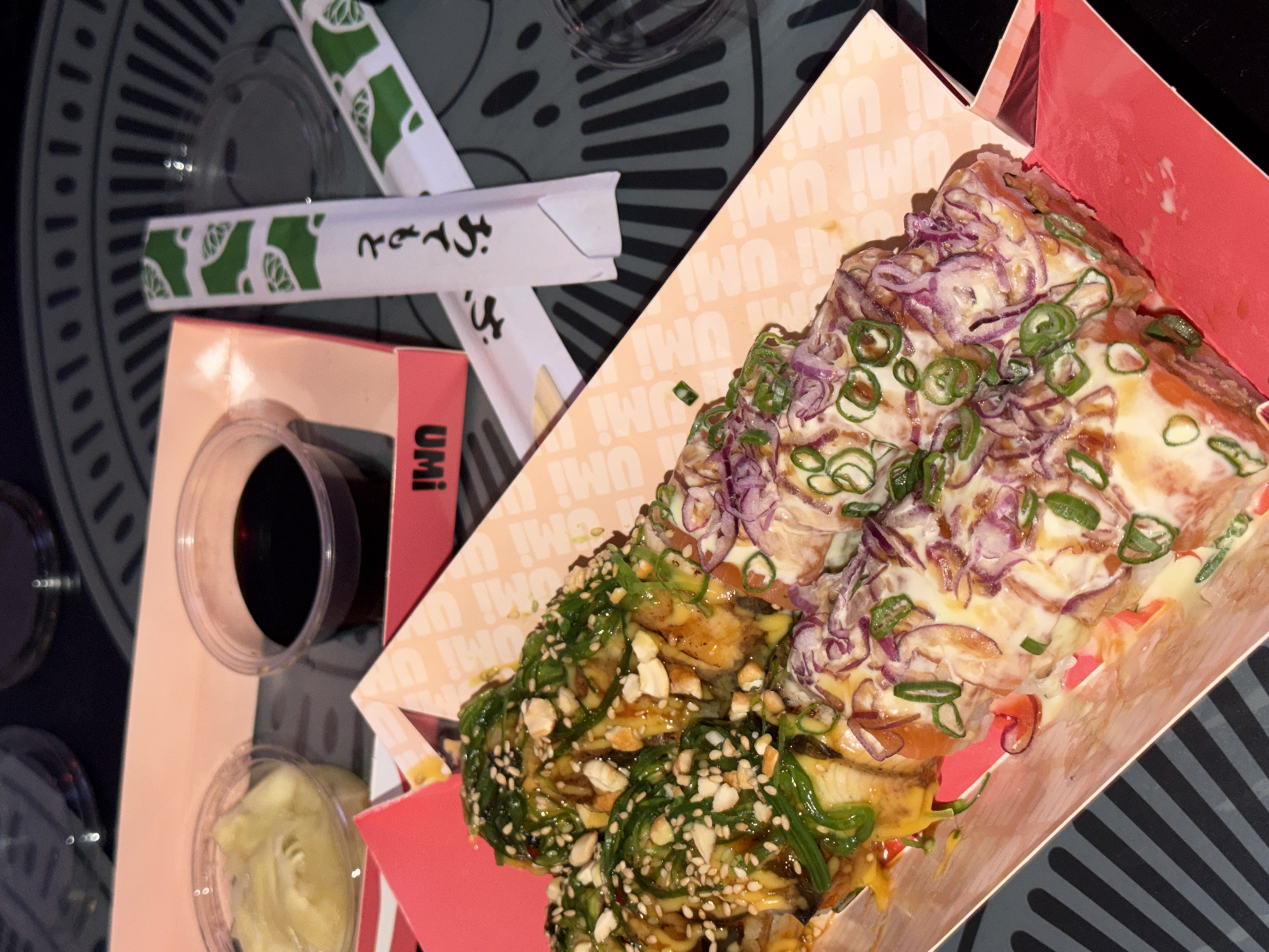 The image shows a close-up shot of food served in an Umi-branded container. The food appears to be sushi or a sushi-inspired dish, topped with seaweed, sesame seeds, chopped green onions, and red onions, and drizzled with sauce. A small container of soy sauce and a container of ginger are visible next to the food. A pair of chopsticks are also present. The surface beneath the food is a dark gray with a radiating striped pattern. The image shows food from Umi restaurant in Belgrade, Serbia.