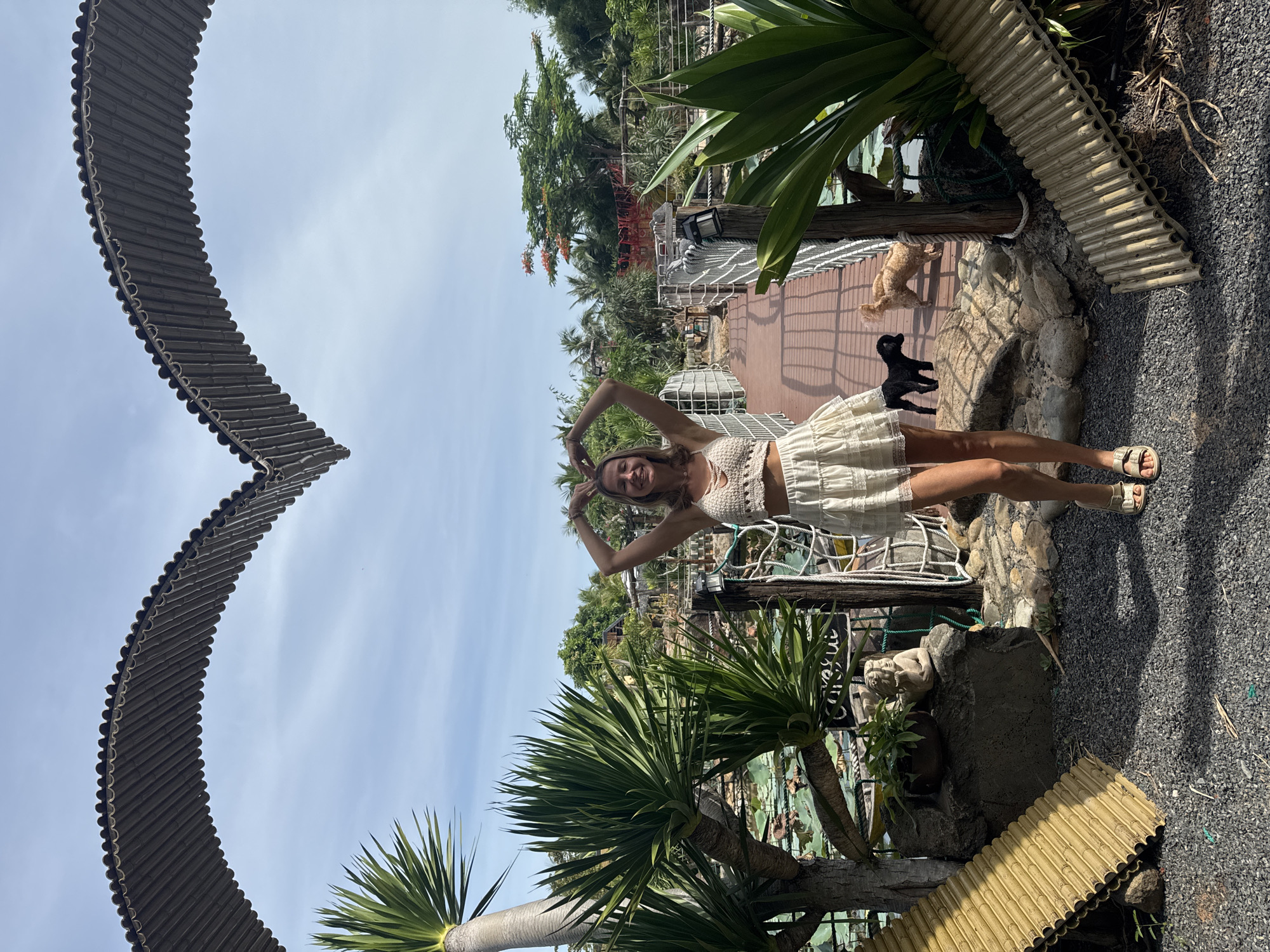 A woman stands within a heart-shaped archway, her hands forming a heart shape above her head. She wears a crocheted crop top and a ruffled skirt. Two dogs, one black and one tan, are visible on a wooden walkway behind her, which leads to a pond with lily pads. The environment is lush and tropical, with palm trees, other greenery, and a clear blue sky. The scene appears to be set in a garden or park, possibly a tourist attraction in Mũi Né, Vietnam.
