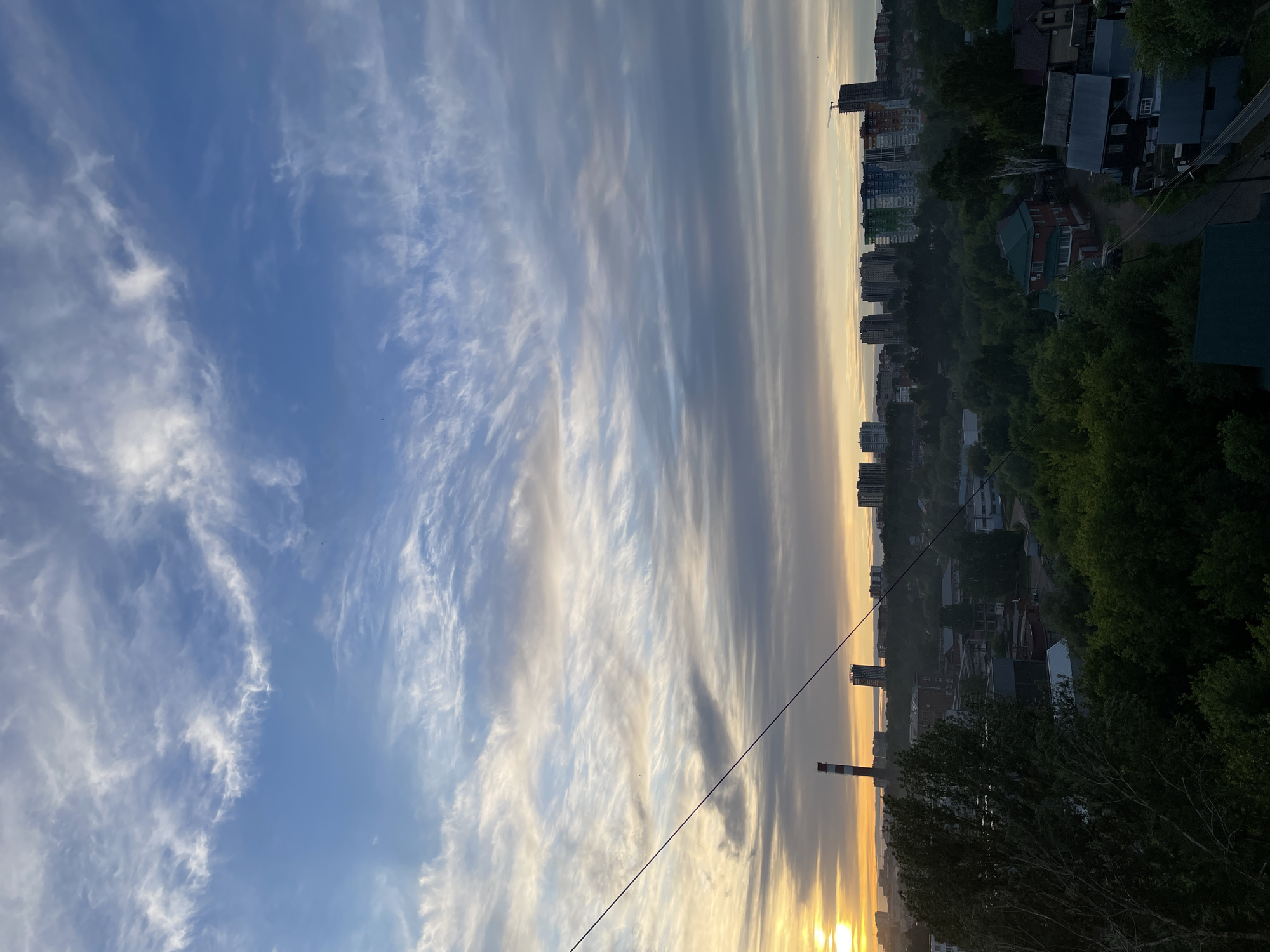 The image captures a view in Izhevsk, Russia. On the right, a cityscape of buildings interspersed with trees stretches into the distance. In the foreground, foliage partially obscures the view. A tall chimney with red and white markings is visible among the buildings. The sky above is filled with elongated cloud formations, with shades of blue and white dominating. Towards the horizon, a gradient of yellow and orange suggests either sunrise or sunset. A power line bisects the lower portion of the image.
