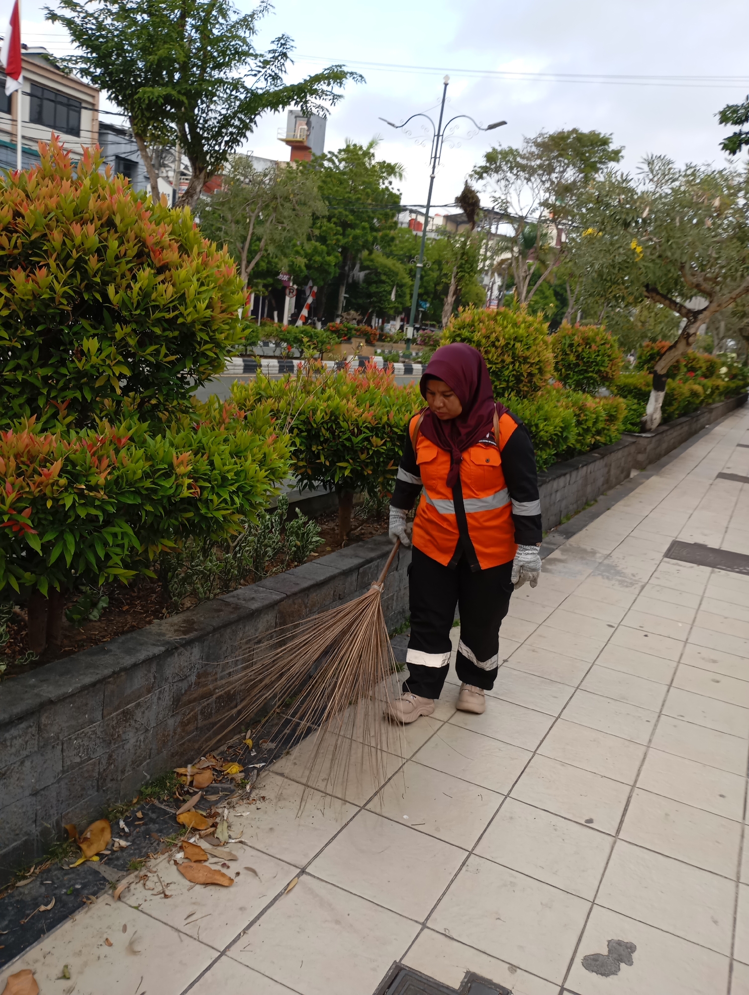 In Balikpapan, Indonesia, a woman wearing a maroon hijab and an orange safety vest over black work pants with reflective stripes is sweeping a tiled sidewalk. She is holding a broom made of natural fibers and is actively sweeping fallen leaves and debris from the edge of a planter box onto the sidewalk. The planter box contains dense, green shrubs with red-tipped new growth. Behind the planter, a road with a black and white striped curb is visible, alongside trees and buildings. The sky is overcast. The overall scene depicts a street cleaning activity in an urban environment.