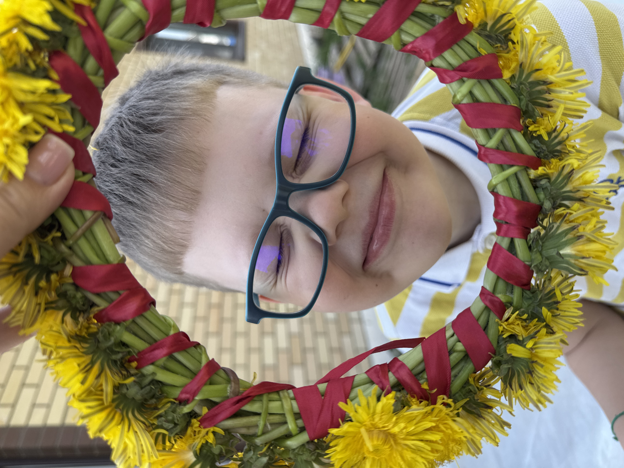 A person wearing glasses is seen through a circular wreath of dandelions and red ribbon, held by a hand with manicured nails. The location is Ферма, Russia, discernible by the visible text on the image. The person, a fair-skinned child, wears a yellow and white striped shirt. The wreath is made of green stems and bright yellow dandelion flower heads. The view is from a low angle, looking upwards. The background shows a building exterior.
