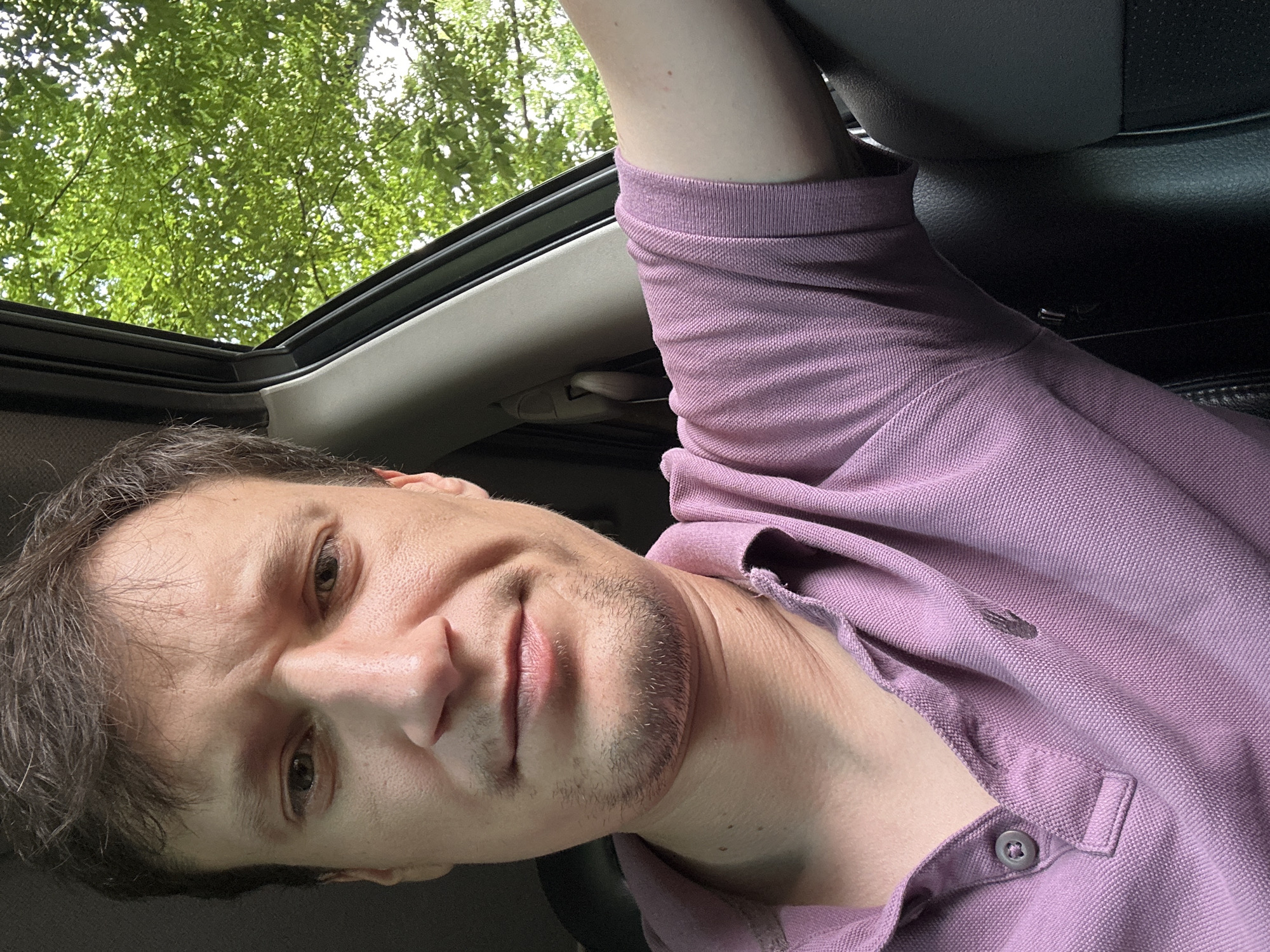 In the city of Shatava, Ukraine, a close-up selfie of a man is captured from the chest up, seated inside a vehicle. The man, with short brown hair and a hint of stubble, wears a lavender polo shirt. His expression is a slight grimace, with furrowed brows and a subtle frown. The background visible through the car window reveals lush green trees and foliage, suggesting an outdoor, possibly rural or park-like setting within Shatava. The interior of the car is dimly lit, with the gray leather of the car seat visible on the right side of the frame. The scene appears to be taken during daylight hours, with dappled light filtering through the leaves.