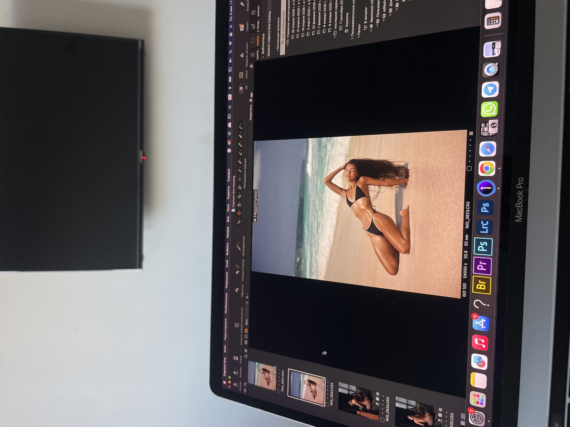 The image shows a close-up view of a MacBook Pro screen displaying the Capture One photo editing software. The software is open to a photo of a woman in a black bikini posing on a beach with turquoise water and white sand. Below the main photo, there is a filmstrip containing previews of other images. Various software icons are visible in the dock, including Adobe Photoshop, Capture One, and a browser. The screen's display settings at the bottom indicate ISO 100, 1/4000 second shutter speed, f/2.8 aperture, and 50mm lens. The MacBook is situated next to a black television. The location in Kazan, Russia is not determinable from the image.