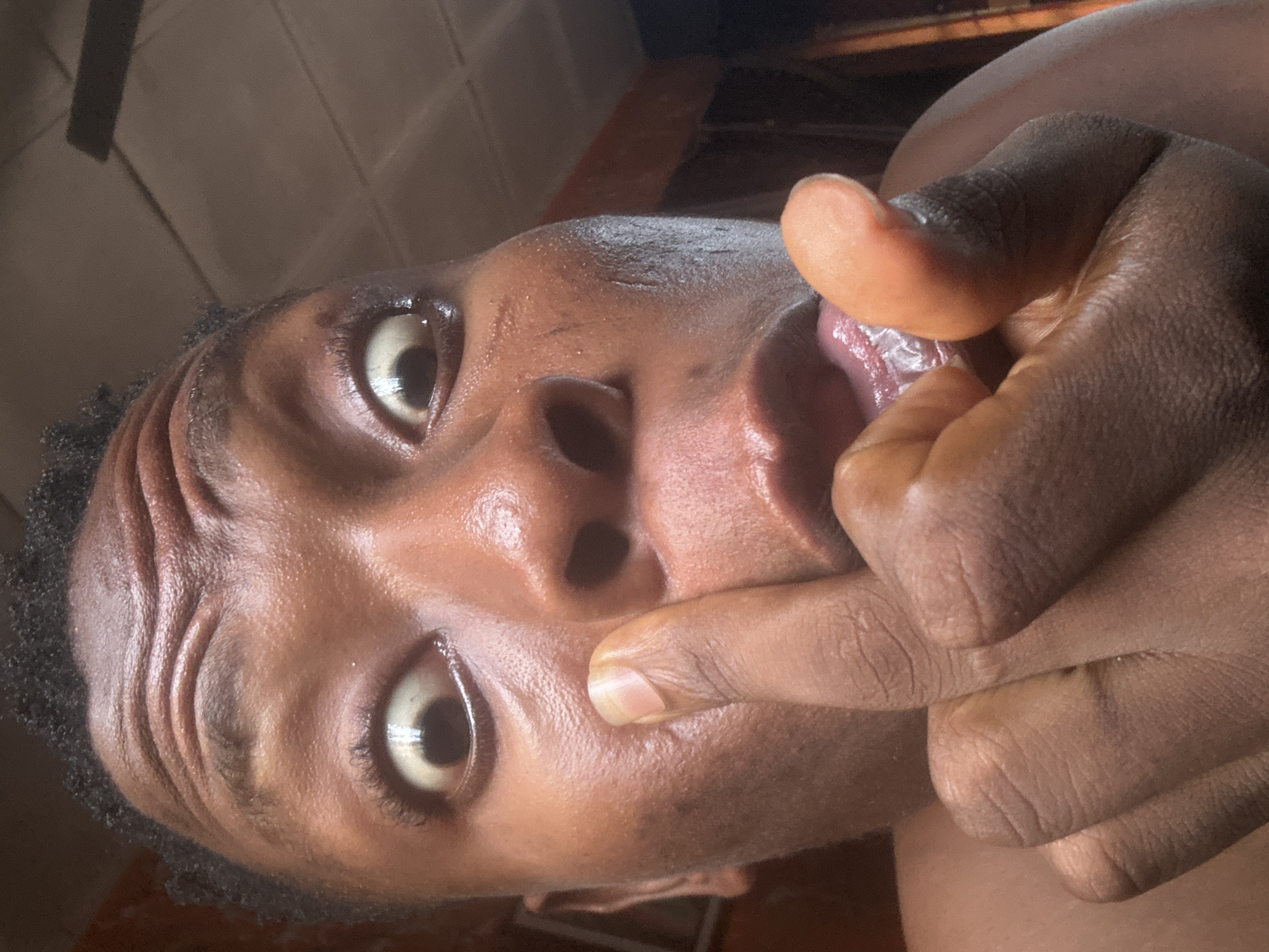 A close-up, selfie-style shot captures a person's face, with their right index finger inserted into their left nostril. The subject's eyes are wide open, with their gaze directed towards the camera. Their mouth is slightly open, revealing their tongue. The background appears to be an indoor setting, possibly a room in a residential building in Ikorodu, Nigeria, indicated by the presence of a ceiling fan and what looks like a tiled ceiling. The lighting suggests it is daytime, with the light source coming from above and slightly to the right. The skin tone of the subject is dark brown, and their hair is short and black. The image focuses intensely on the facial expression and the action being performed, creating an intimate and somewhat startling composition.