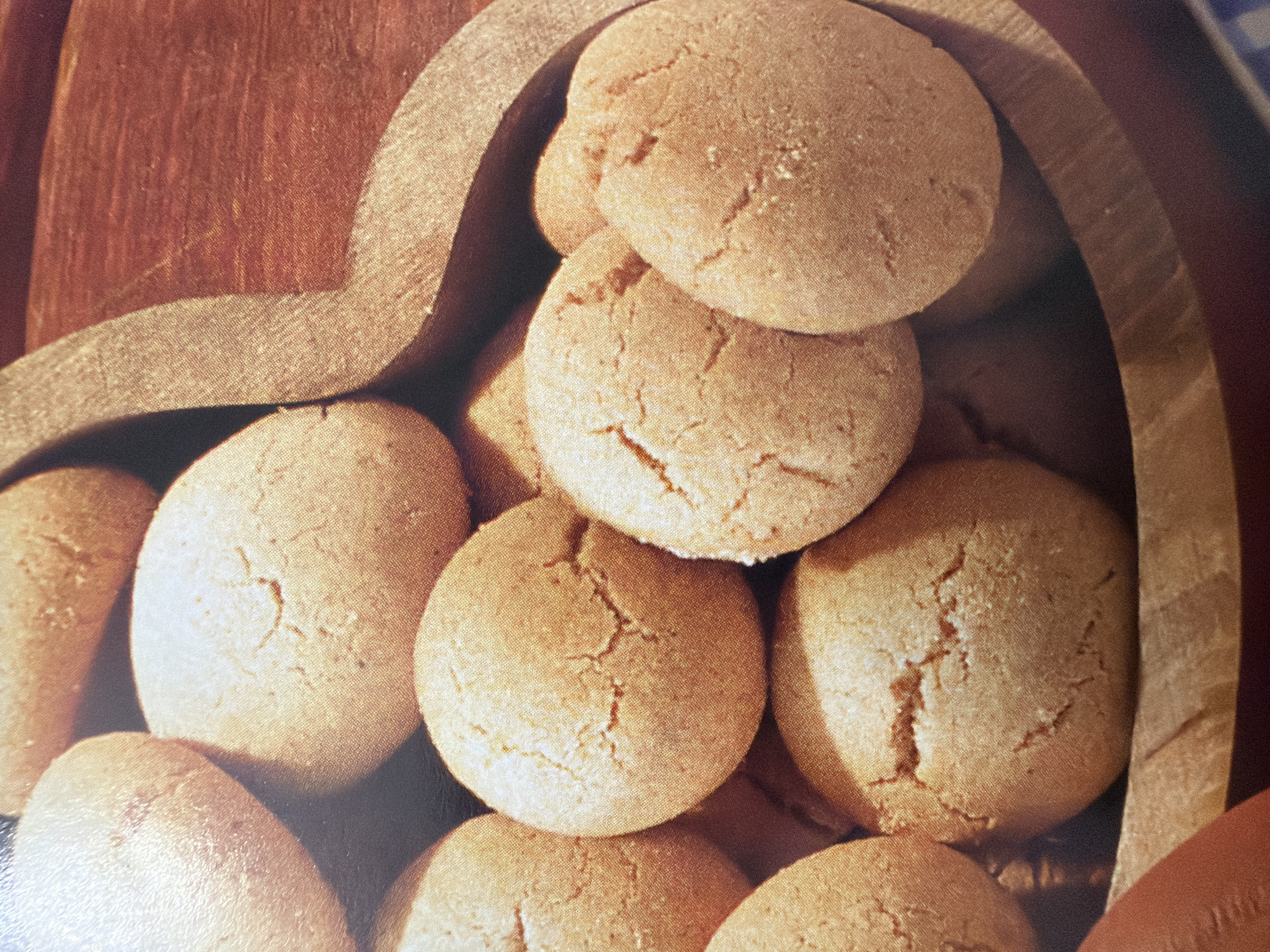 An overhead, close-up, studio shot captures a collection of golden brown, spherical cookies with cracked surfaces, nestled within a carved, heart-shaped wooden bowl. The cookies are uniformly baked, exhibiting a soft texture. The wooden bowl is deep and polished, with visible wood grain and a natural, rustic finish. The lighting is soft and diffused, highlighting the textures and colors of the cookies and bowl. The background is dark and out of focus, further emphasizing the food items. The image appears to be a still-life arrangement for promotional or culinary purposes. The scene is set in the city of Unknown, Unknown.