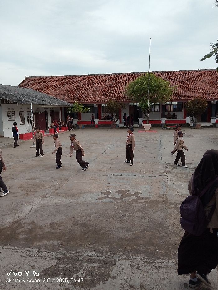 The image displays an outdoor school courtyard in Wanguk, Indonesia, captured at 06:47 on October 3, 2025, under an overcast sky. In the background, a long, single-story school building features a red tiled roof, white pillars, and a red-painted lower wall section; multiple students are visible seated on ledges or standing under its porch. To the left, a smaller white building with a dark, possibly corrugated roof stands. A tall, white flagpole is positioned centrally in the background, with a green tree immediately behind it. The ground is a worn concrete or asphalt surface with various stains and cracks.
Approximately 15-20 individuals, primarily children identified as students, are present. The majority wear uniforms consisting of light brown shirts and dark trousers or skirts, some with dark hats, resembling Indonesian school or scout uniforms. In the mid-ground, at least six male students are observed standing or walking across the courtyard; some are in motion towards the left, others are stationary. A female student is partially visible in the right foreground, seen from behind, wearing a black hijab, a light brown uniform shirt, a dark skirt, and a purple backpack. One male individual in a blue and red shirt stands near the smaller building on the far left.
