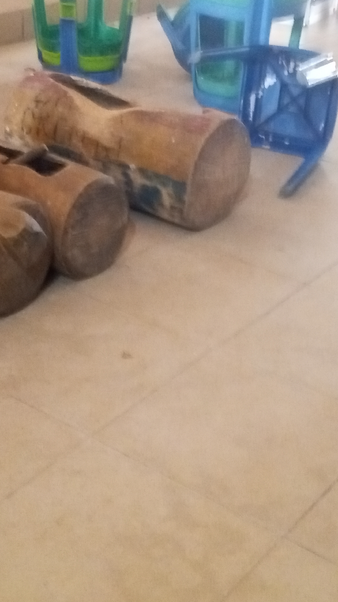 A blurred, close-up, overhead shot shows wooden mallets resting on a tiled floor. In the background, blue and green plastic chairs are stacked. The scene is dimly lit, and the focus is on the mallets, which appear worn and possibly old. The overall impression is one of disarray or storage.