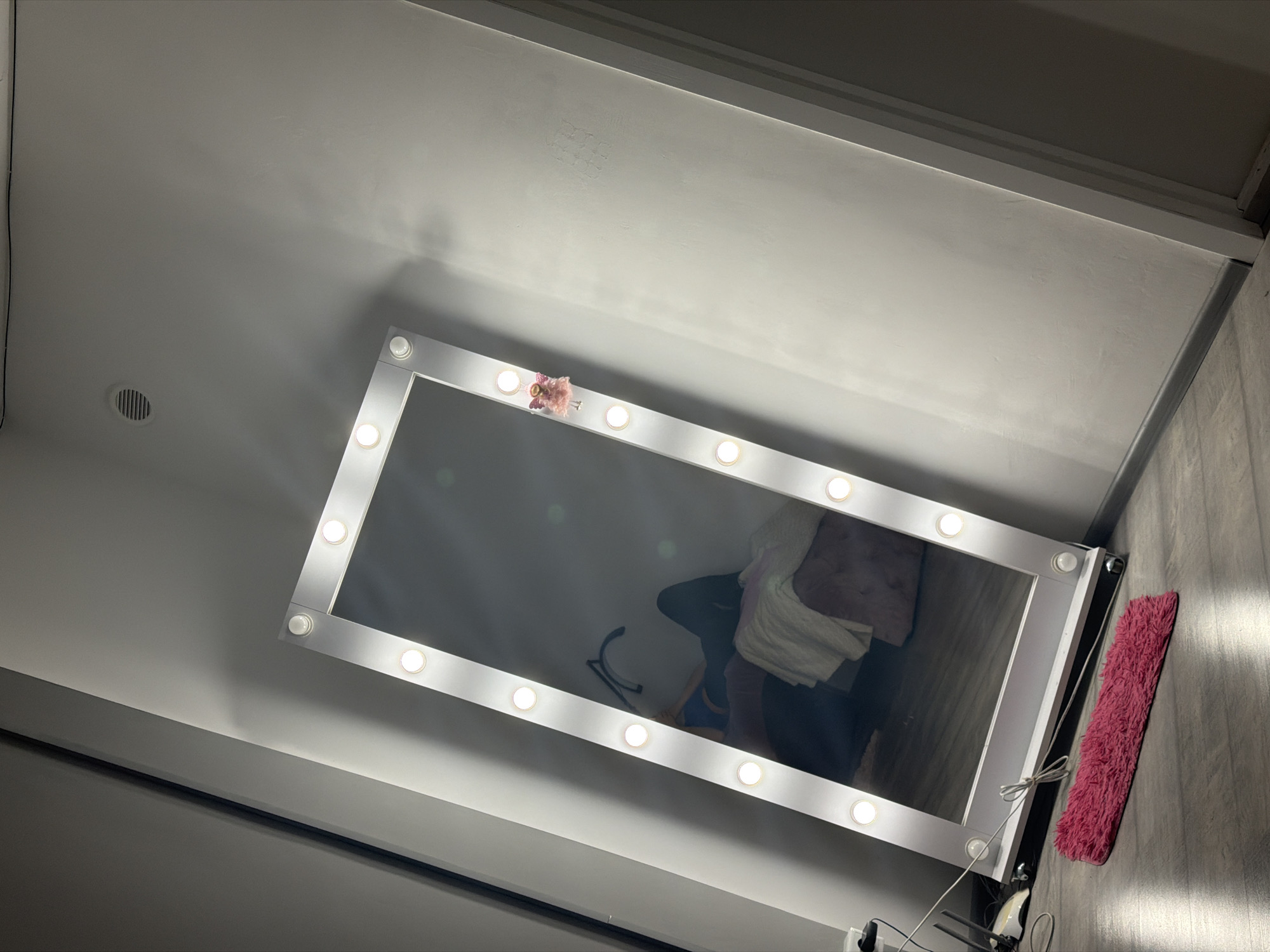 In a room with light gray walls and a wooden floor, a large, rectangular vanity mirror with integrated LED lights stands on wheels. The mirror reflects a person lying down on a pink ottoman, wearing a blue top and black pants. A white blanket is draped over the ottoman. A small, pink decorative angel figure is attached to the side of the mirror frame. A pink shag rug lies on the floor in front of the mirror. To the left of the mirror, a white router and some cables are visible near the wall.