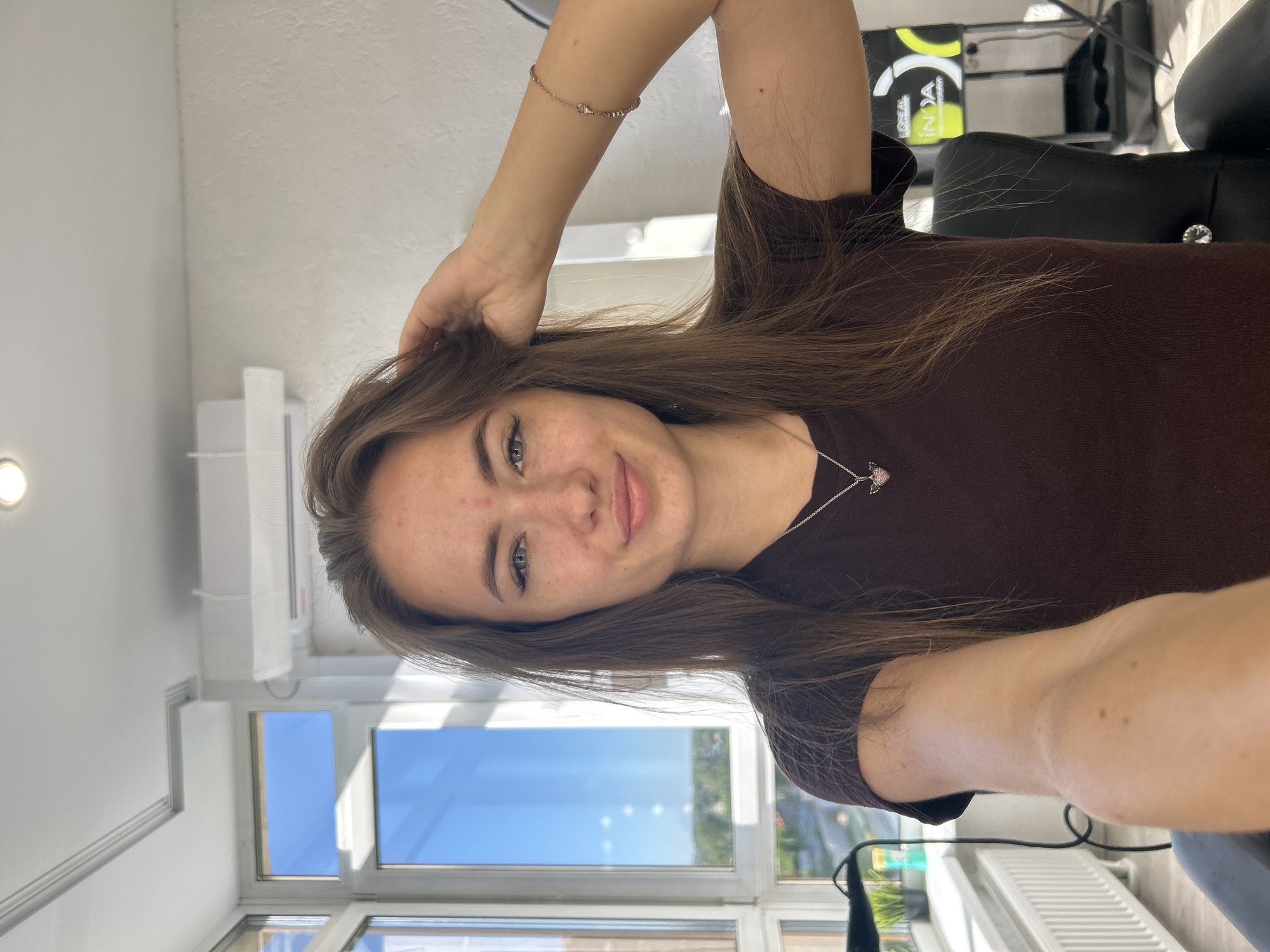 A young Caucasian woman with long brown hair is pictured from the chest up, facing the camera with her right hand on the back of her head. She is wearing a dark brown t-shirt and a silver necklace with a pink heart-shaped pendant. The background features a large window with a view of the sky and trees, and an air conditioner unit mounted on the wall. To the right, a black chair and a hair salon display with L'Oréal branding are visible. The woman has light skin with some freckles and is smiling slightly.